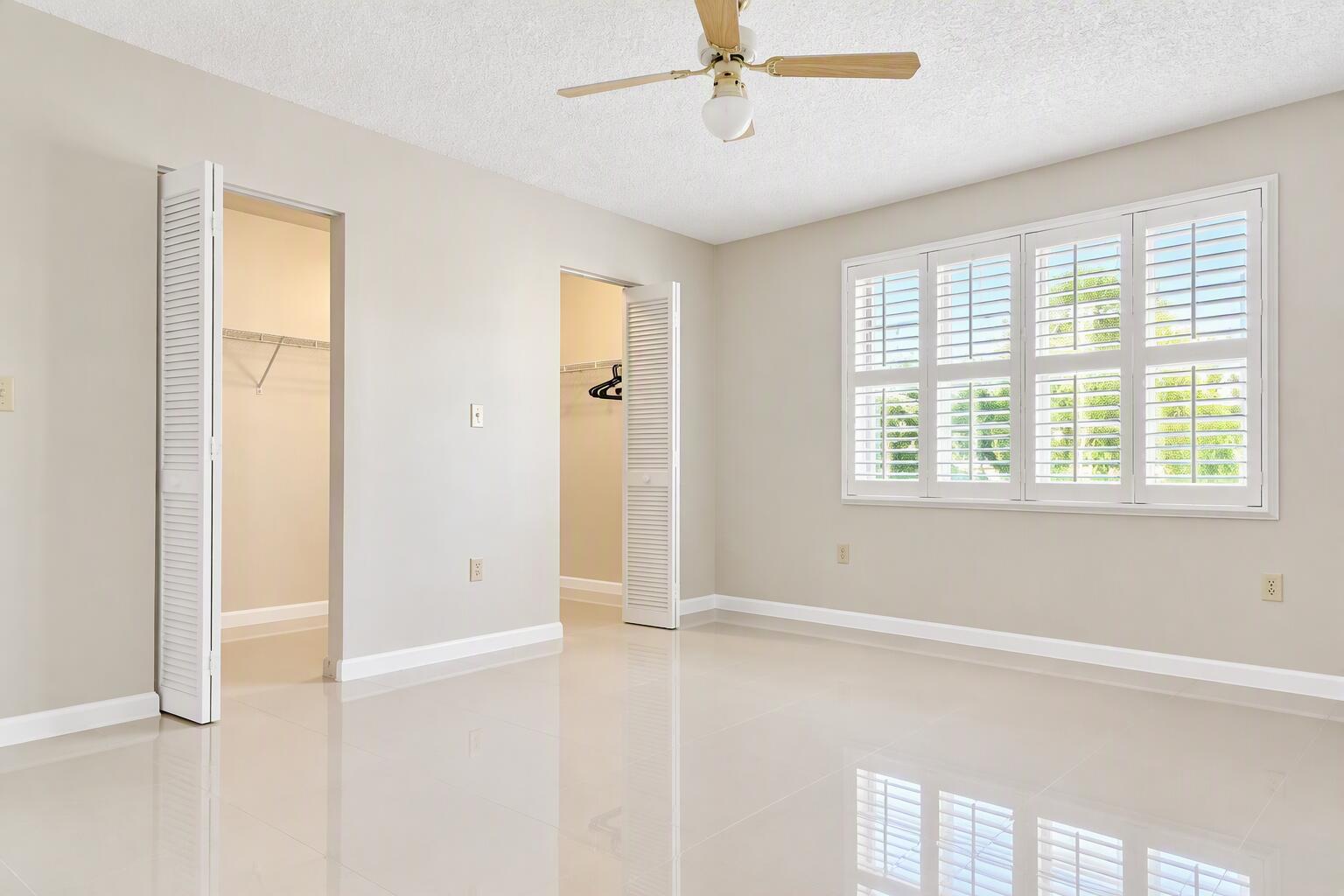 Neutral-toned interior featuring polished tile flooring, window shutters, dual closet entries with louvered doors, and a ceiling fan with light fixture