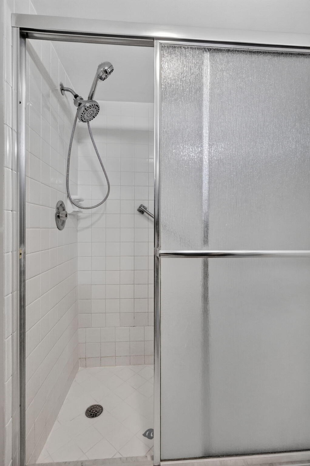Shower enclosure featuring white tile walls, chrome-framed sliding textured glass doors, and a handheld shower fixture