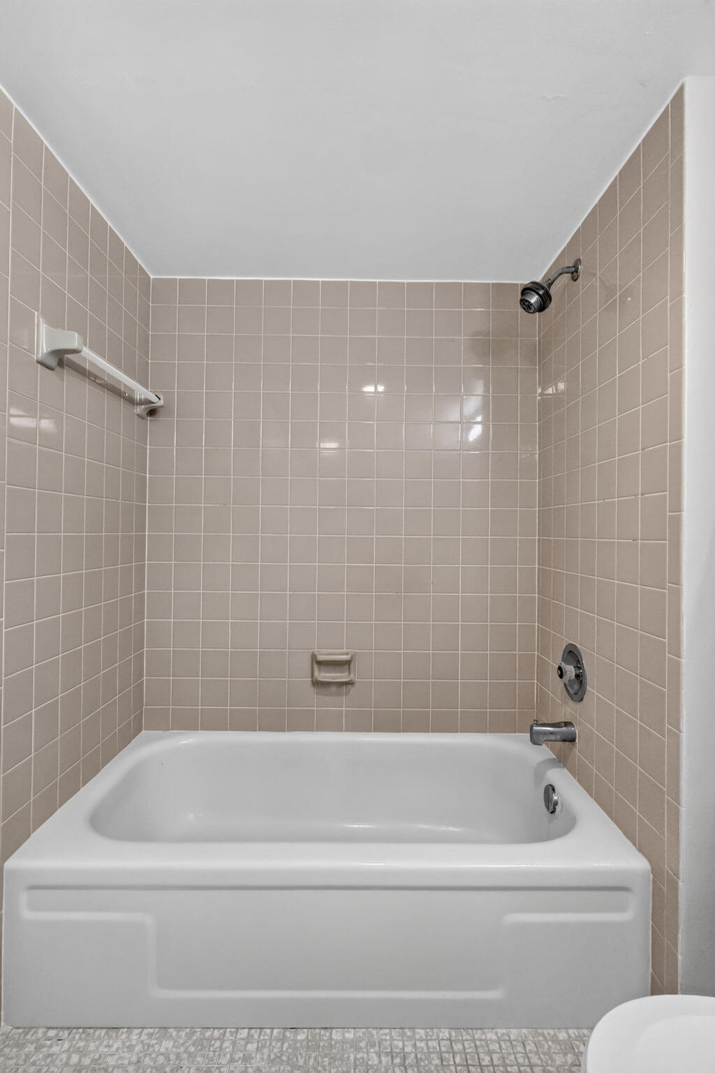 Bathtub surround featuring light-toned square wall tiles, integrated soap dish, chrome shower head, and chrome faucet hardware