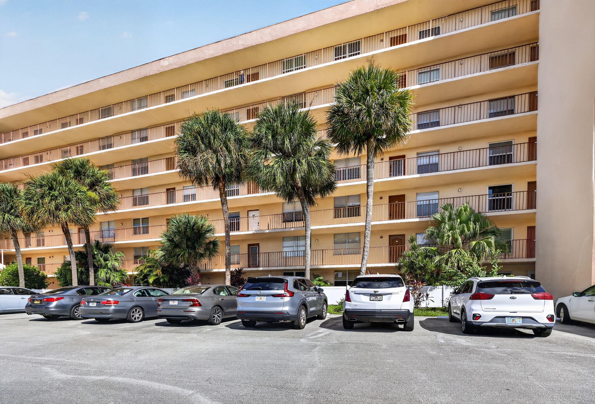 Multi-story building exterior featuring a curved facade, private balconies with metal railings, and mature palm tree landscaping