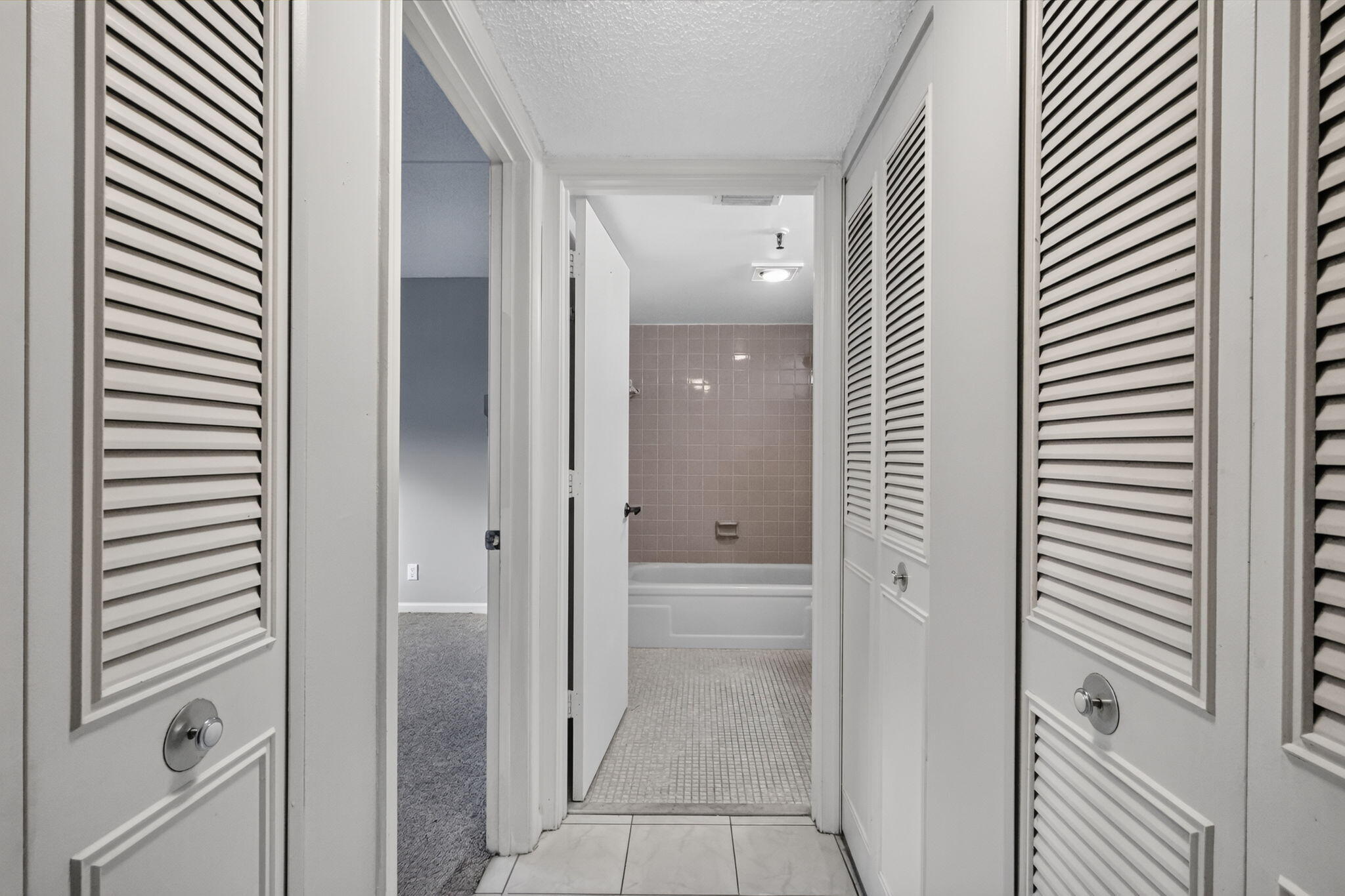 Hallway featuring dual floor-to-ceiling closets with white louvered doors, tile flooring, and a bathroom containing a tiled shower surround