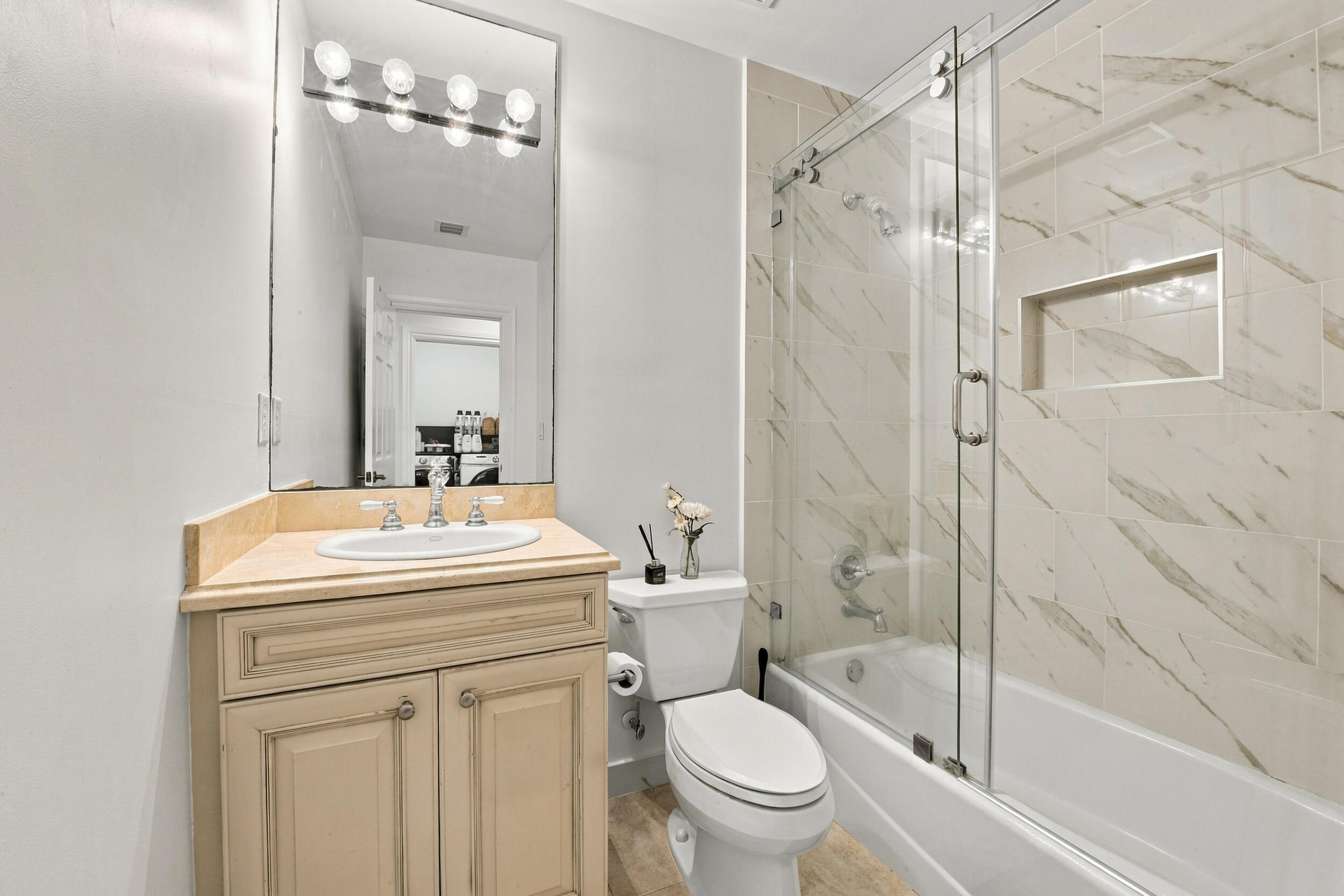 Bathroom featuring a vanity with a light-finish cabinet and an integrated oval sink, a frameless sliding glass shower door, and a shower enclosure tiled with a stone-finish surround featuring an integrated storage niche