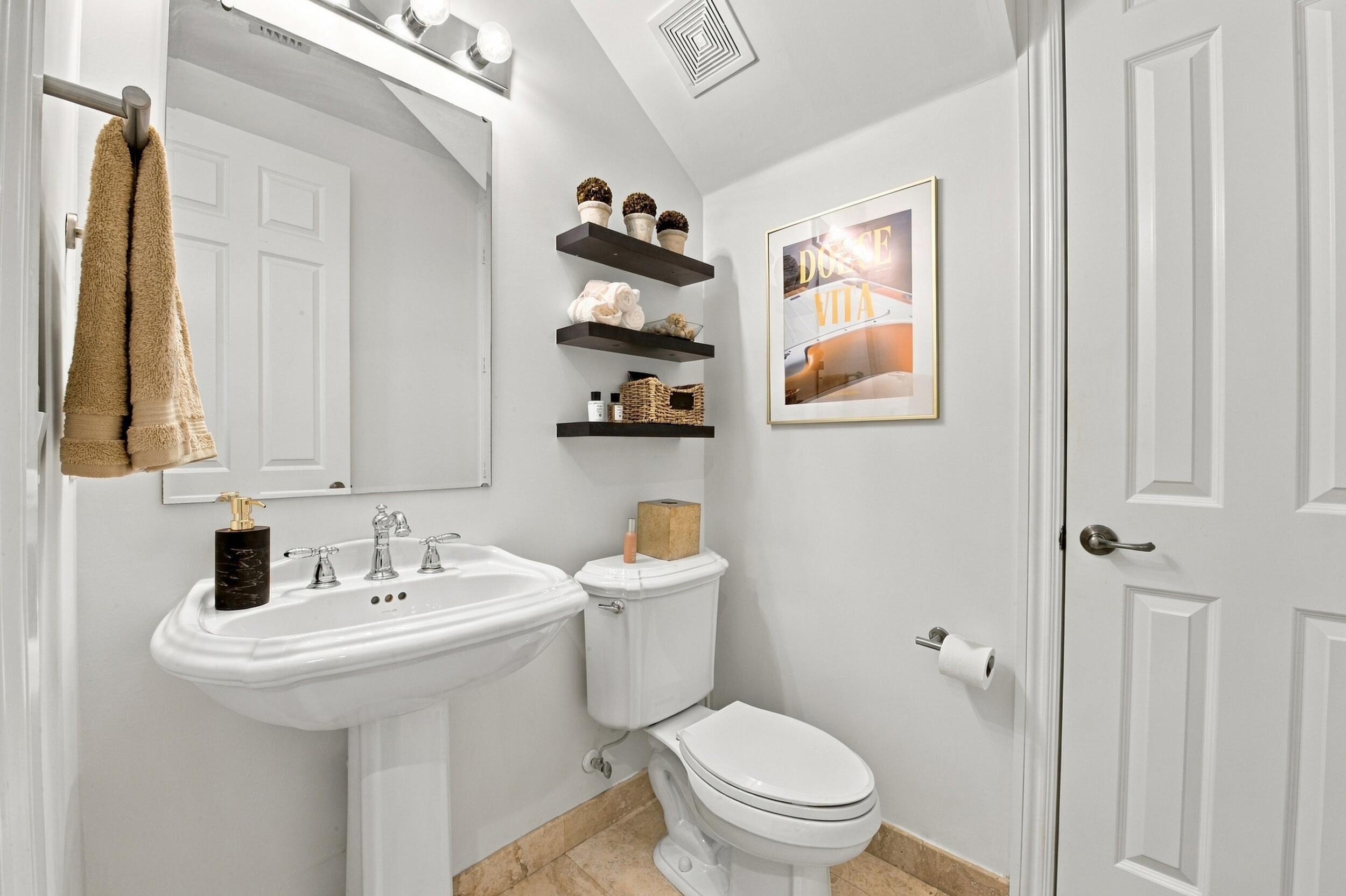 Pedestal sink featuring a polished chrome faucet, wall-mounted light fixture, mirror, three built-in floating shelves, and stone-finish flooring
