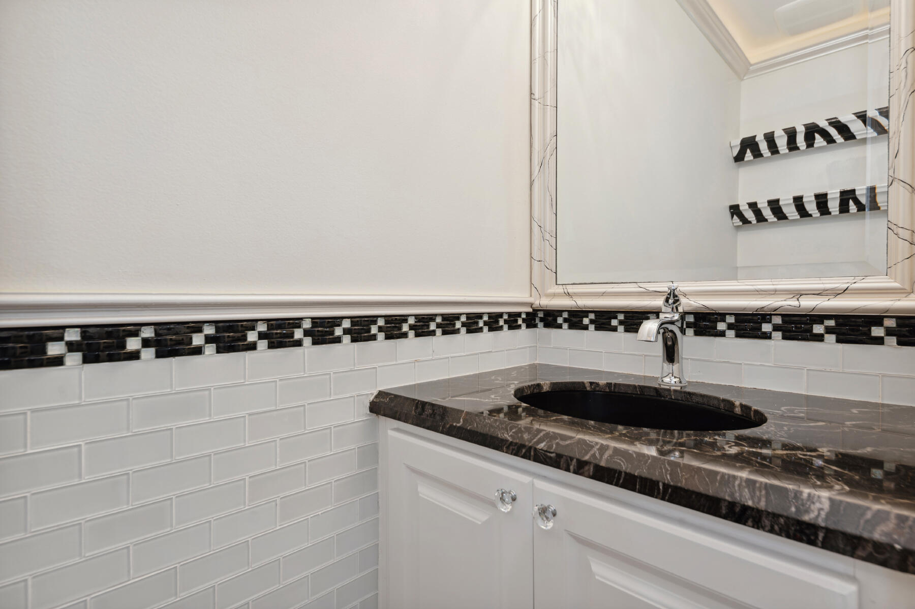 Vanity featuring a dark stone countertop, undermount oval sink, polished chrome faucet, white cabinetry with crystal-style hardware, and a subway tile backsplash with a decorative checkered border