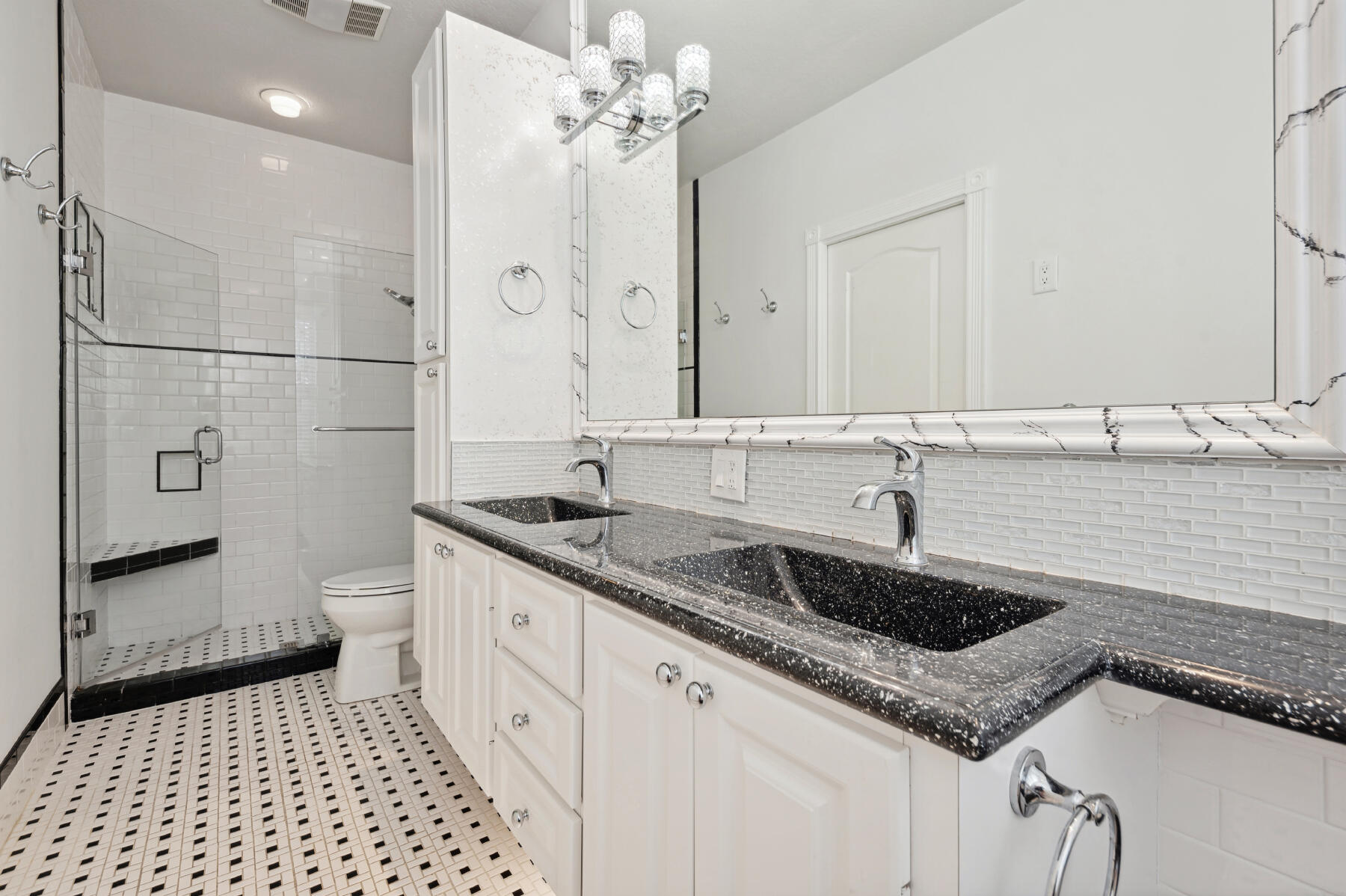 Double vanity with dark speckled countertop and integrated sinks, white cabinetry, glass-enclosed shower with subway tile surround, and mosaic tile flooring