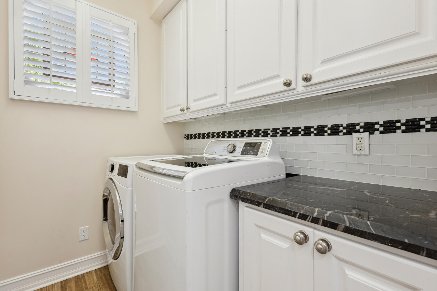 Laundry area featuring white shaker-style cabinetry, dark stone-finish countertops, subway tile backsplash with decorative inset, and plantation shutters