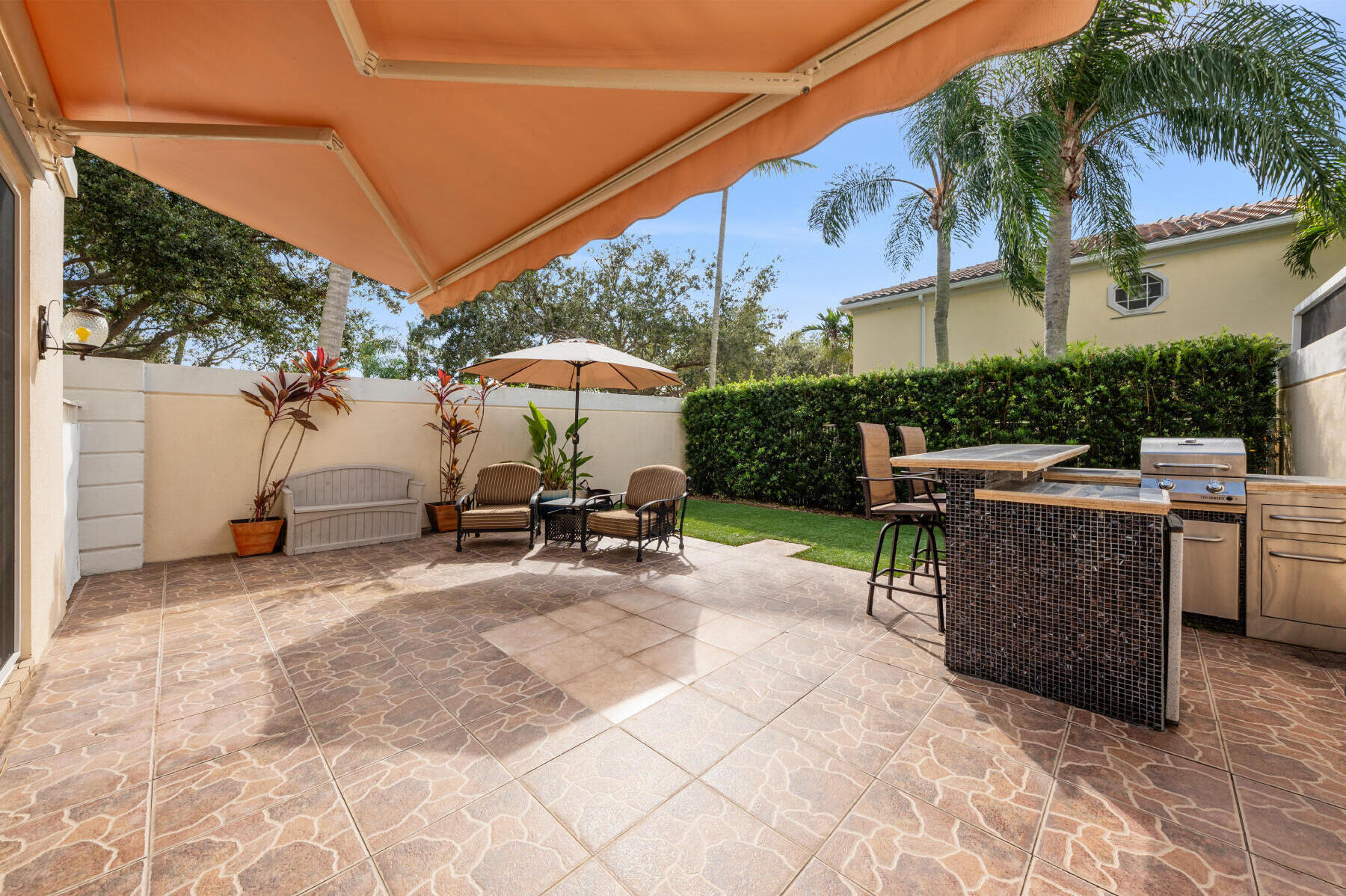Outdoor patio featuring a retractable awning, tiled flooring, and a built-in outdoor kitchen with stainless steel appliances and mosaic tile detailing, bordered by privacy walls and mature landscaping
