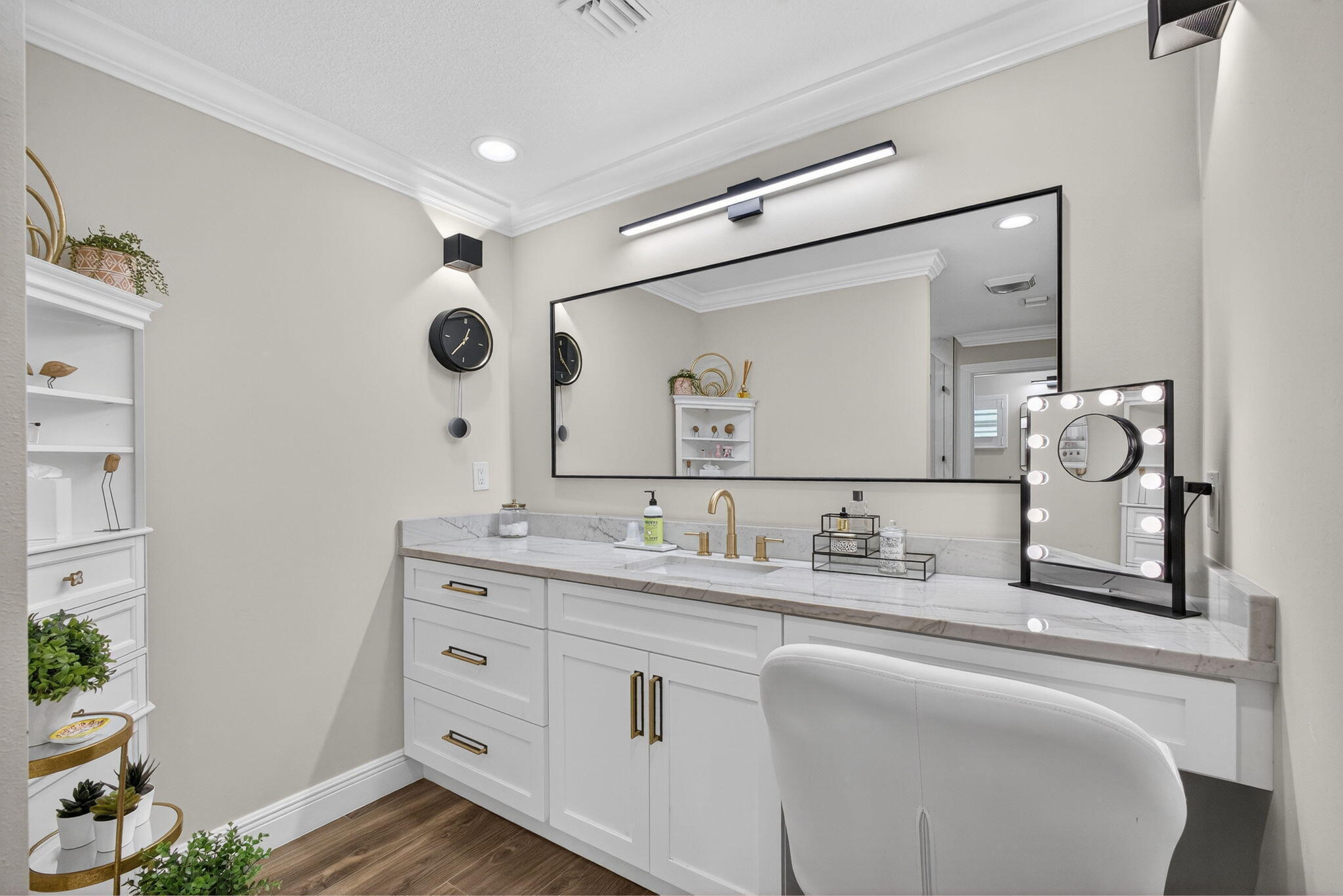 White vanity cabinetry featuring gold-tone hardware, stone-finish countertop with an integrated sink, polished gold-tone faucet, large rectangular mirror with a black frame, and linear LED vanity lighting