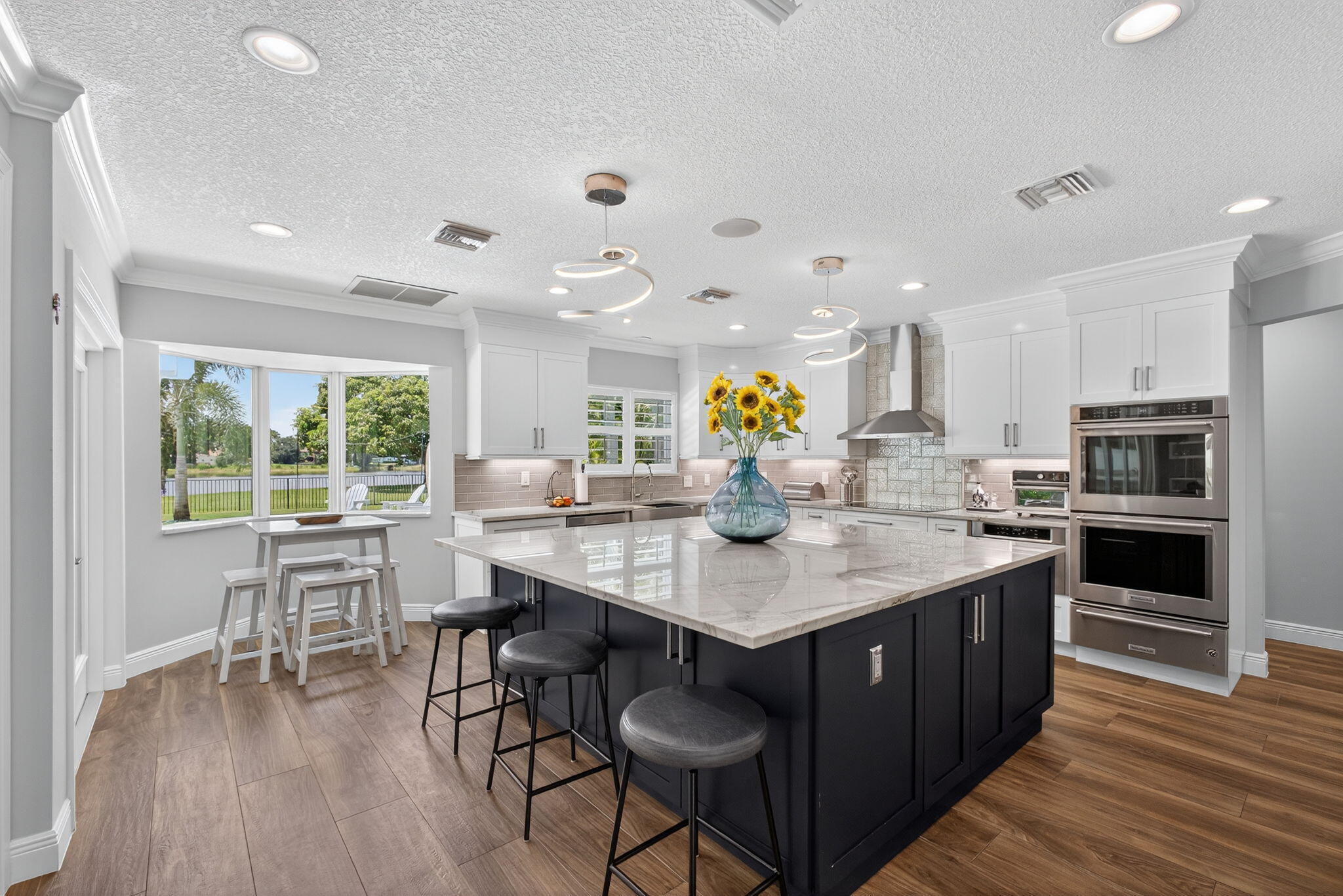 Kitchen featuring a contrasting dark base cabinet island with a stone countertop, white upper cabinetry, built-in stainless steel double ovens, recessed ceiling lighting, and wood-finish flooring