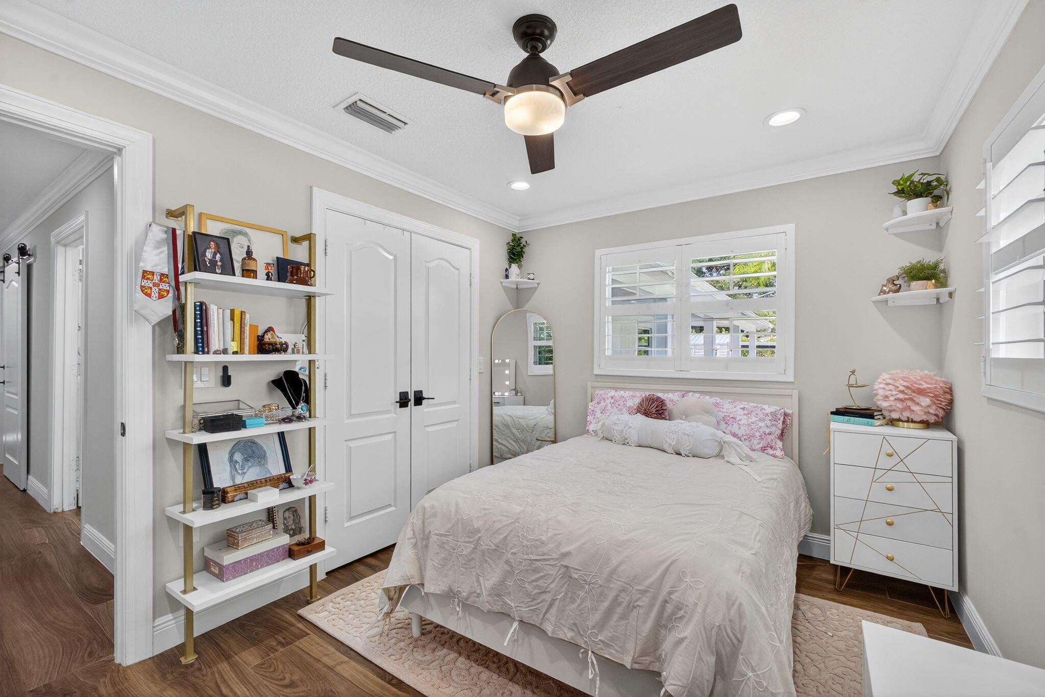 Neutral interior featuring wood-finish flooring, crown molding, double-door closet with matte black hardware, dual-blade ceiling fan with integrated lighting, and white louvered plantation shutters