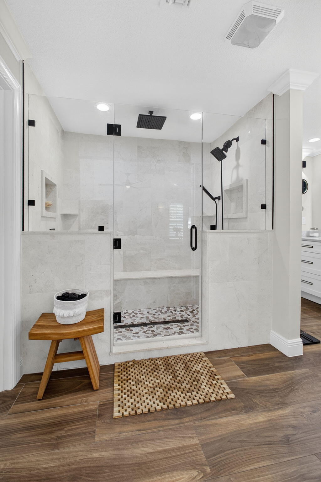 Walk-in shower featuring a frameless glass enclosure, matte black fixtures, integrated shelving, and multi-tone mosaic tile flooring, accented by wood-finish flooring
