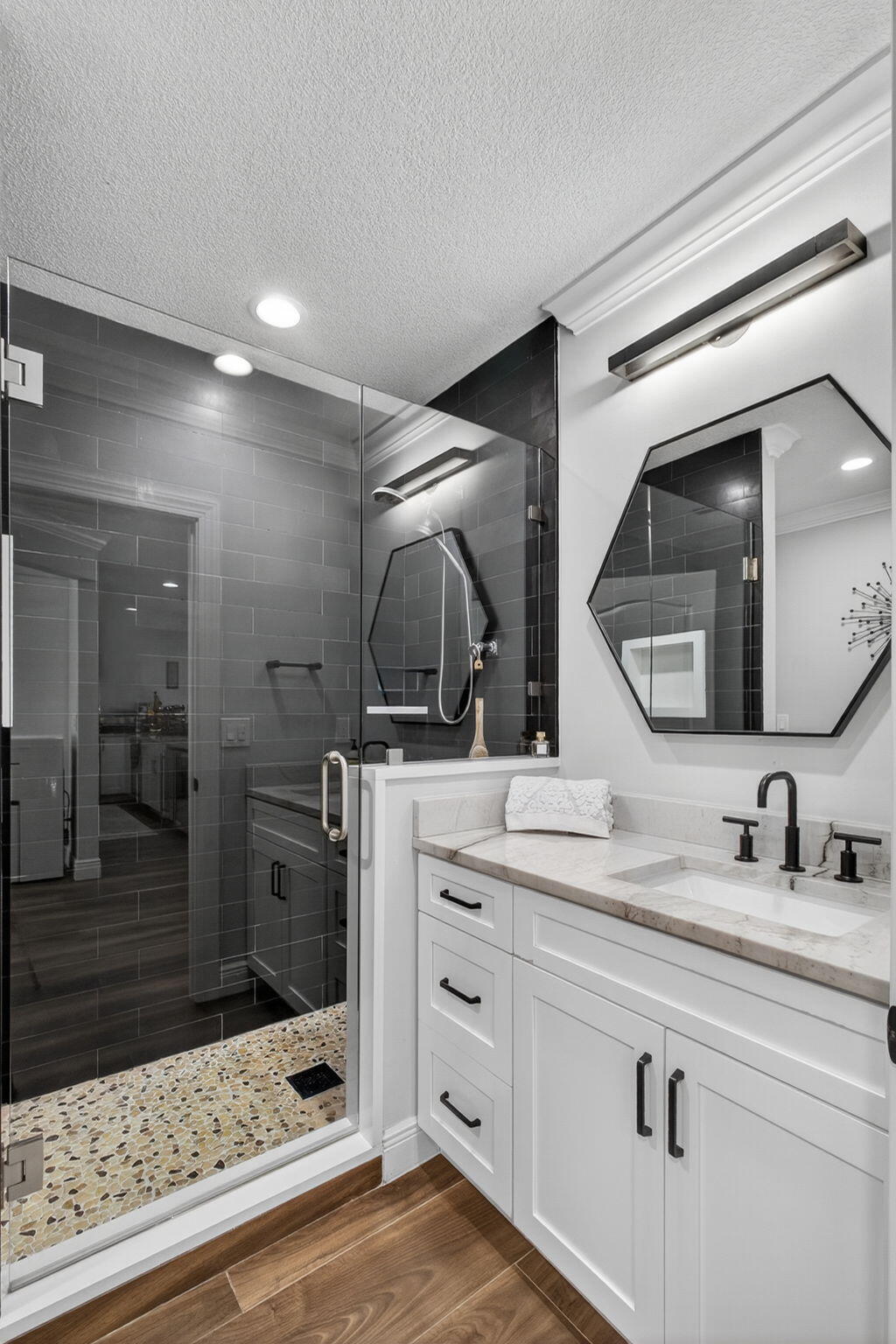White shaker-style vanity with stone countertop, undermount sink, and matte black hardware, paired with a glass-enclosed shower featuring stone flooring, subway tile walls, and a recessed niche