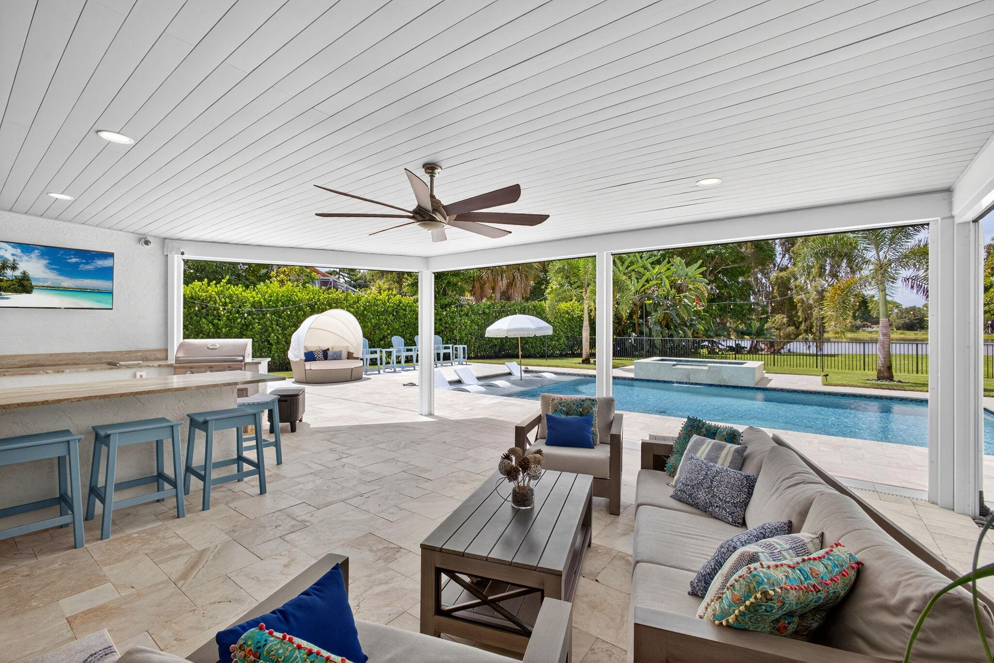 Covered outdoor living space featuring a stone-tile patio, built-in kitchen with stone-top bar, recessed lighting, ceiling fan, and swimming pool with a raised spillover spa