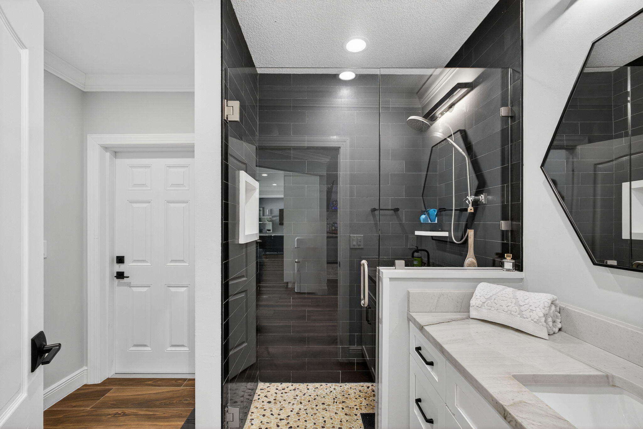 Bathroom featuring a walk-in shower with charcoal subway tile, pebble-stone flooring, frameless glass enclosure, white cabinetry with stone-finish countertop, and a geometric-framed wall mirror