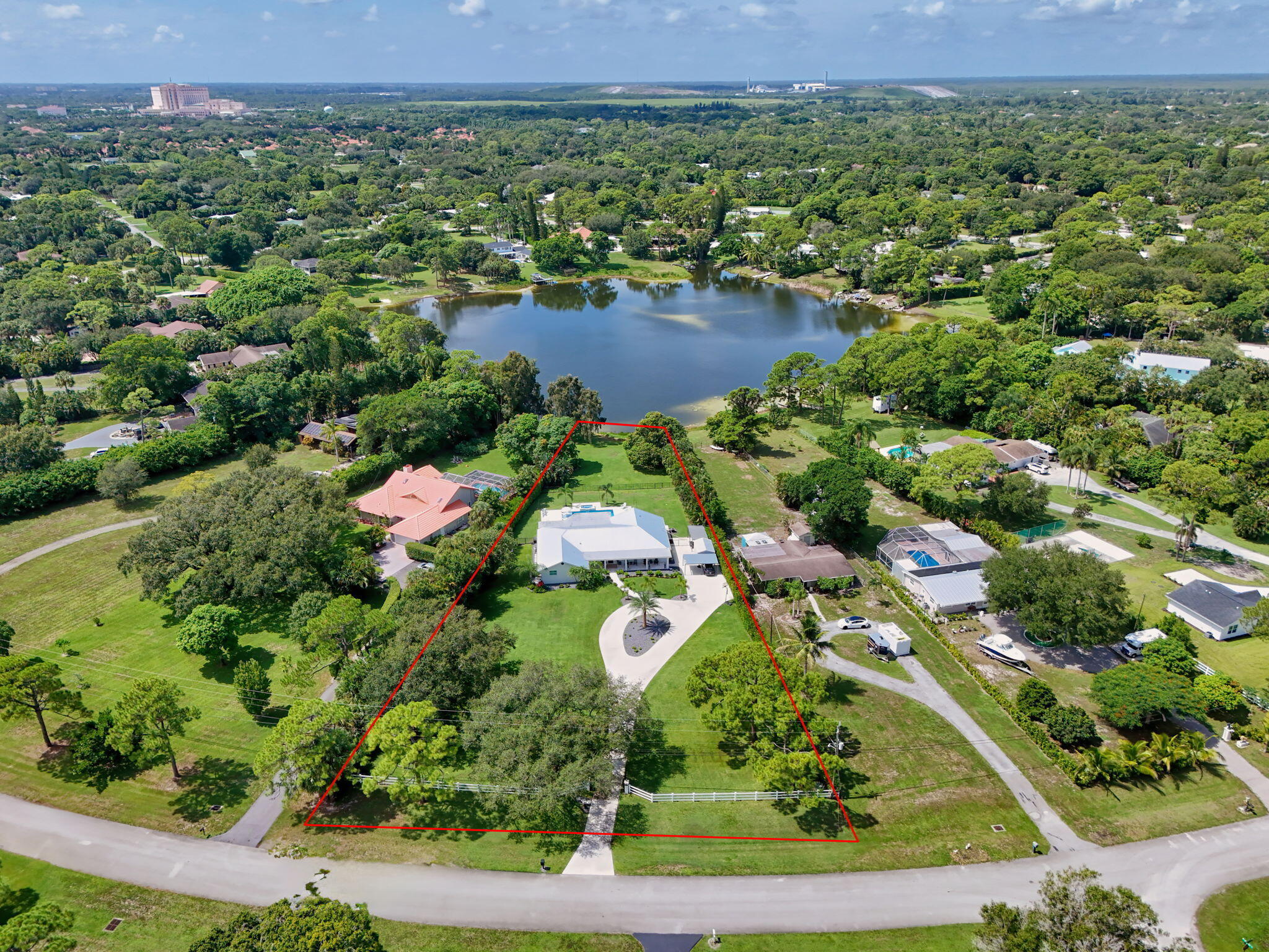 Aerial perspective of a sprawling property featuring a paved circular driveway, white post-and-rail fencing, a manicured lawn, and mature canopy trees situated on a lakefront lot