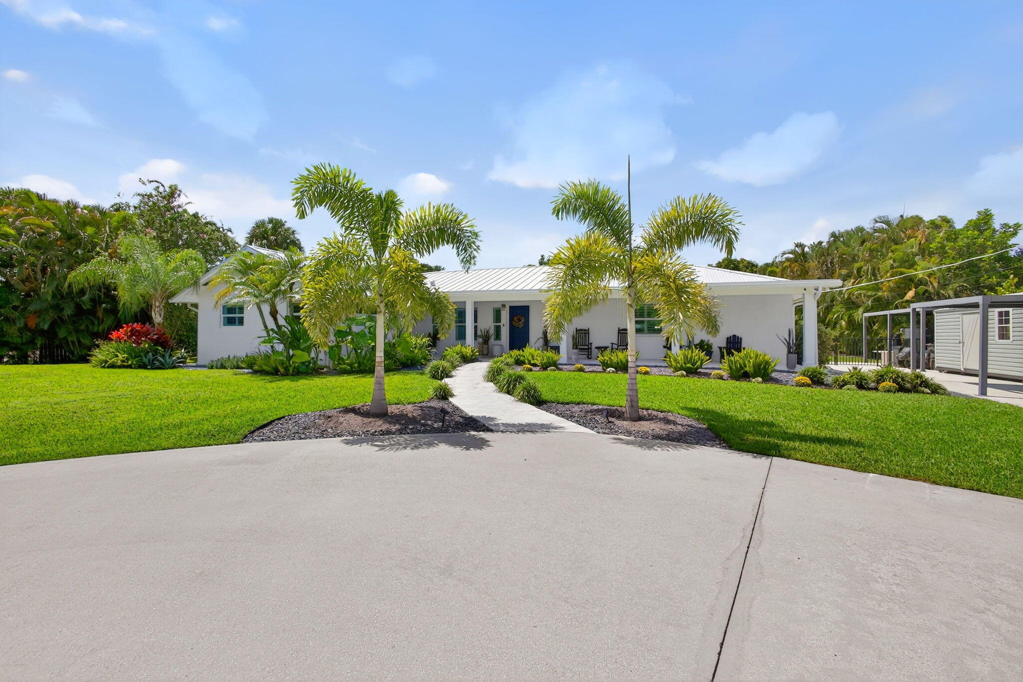 Single-story exterior featuring white masonry walls, a metal roof, a paved walkway, a spacious concrete driveway, and established palm trees