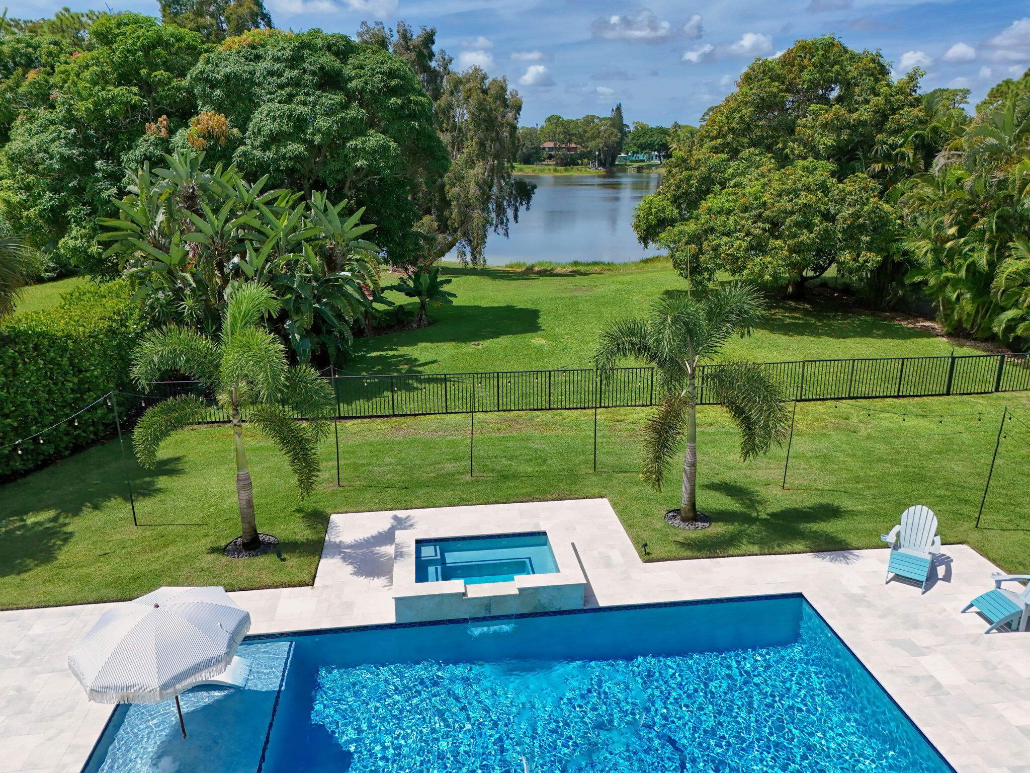 In-ground swimming pool and attached spillover spa featuring light-colored stone decking, set against a backdrop of waterfront scenery and landscaped greenery