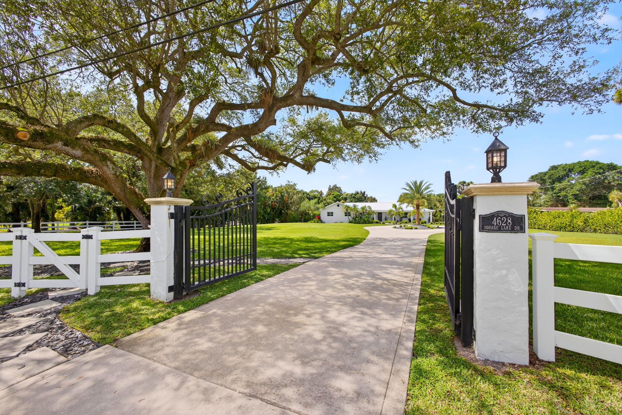 Gated entrance featuring decorative metal fencing, concrete-paved driveway, stucco-finished stone pillars, and mounted lantern-style lighting
