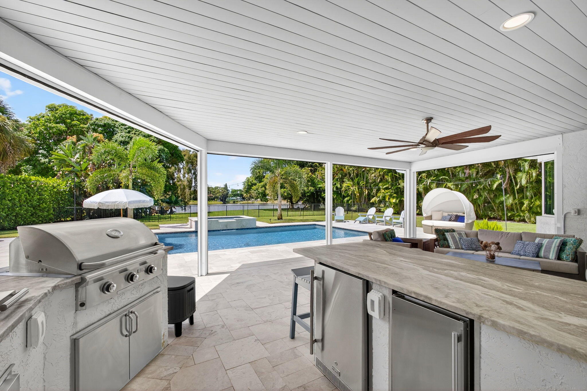Covered patio featuring an outdoor kitchen with stainless steel appliances, stone-finish countertops, tile flooring, and a ceiling fan, opening to a swimming pool with a built-in spa