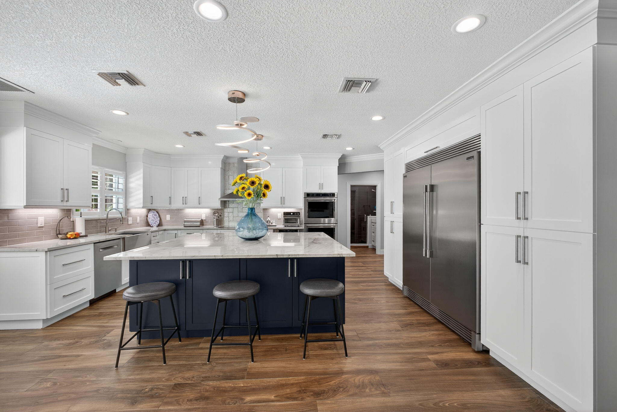 Kitchen featuring custom white cabinetry, a contrasting navy blue center island, stone-finish countertops, stainless steel appliances, and wood-finish flooring