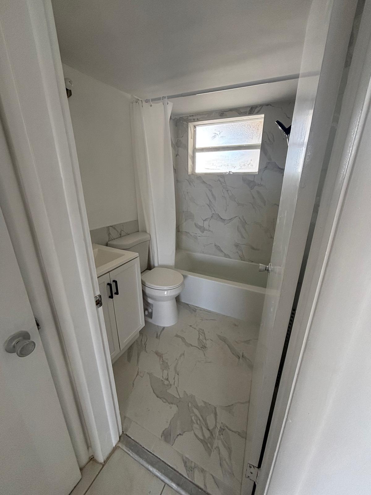 Bathroom featuring stone-finish floor tiling, tub-shower combination with floor-to-ceiling stone-finish wall surrounds, single-basin vanity cabinet with matte black hardware, and an exterior window