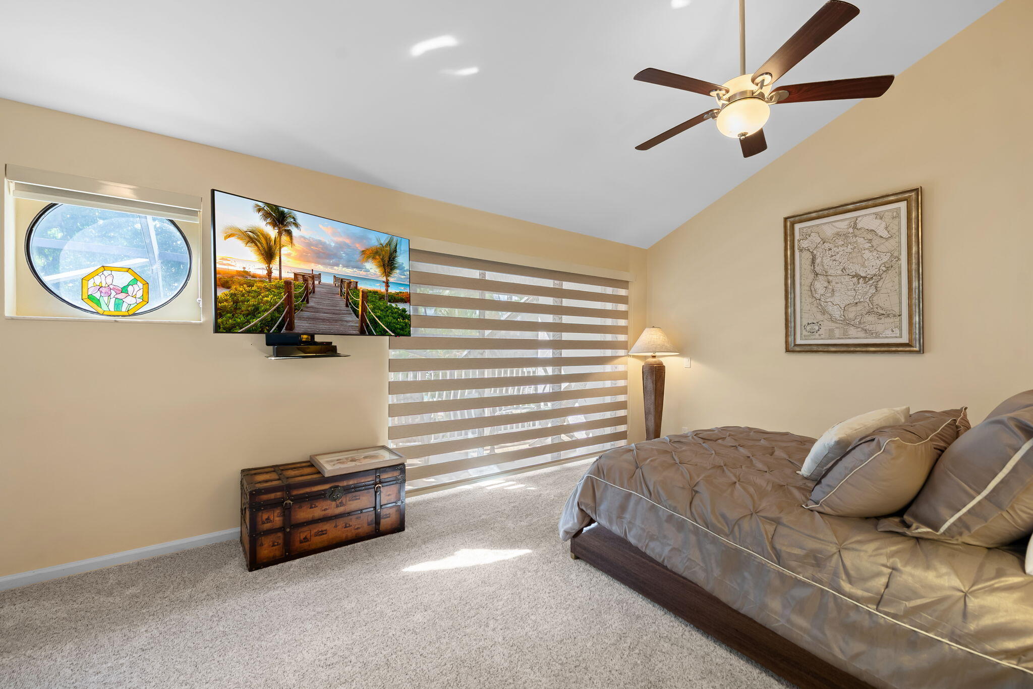 Vaulted ceiling with a central multi-blade ceiling fan, circular accent window featuring decorative glass, and large dual-layer horizontal blinds