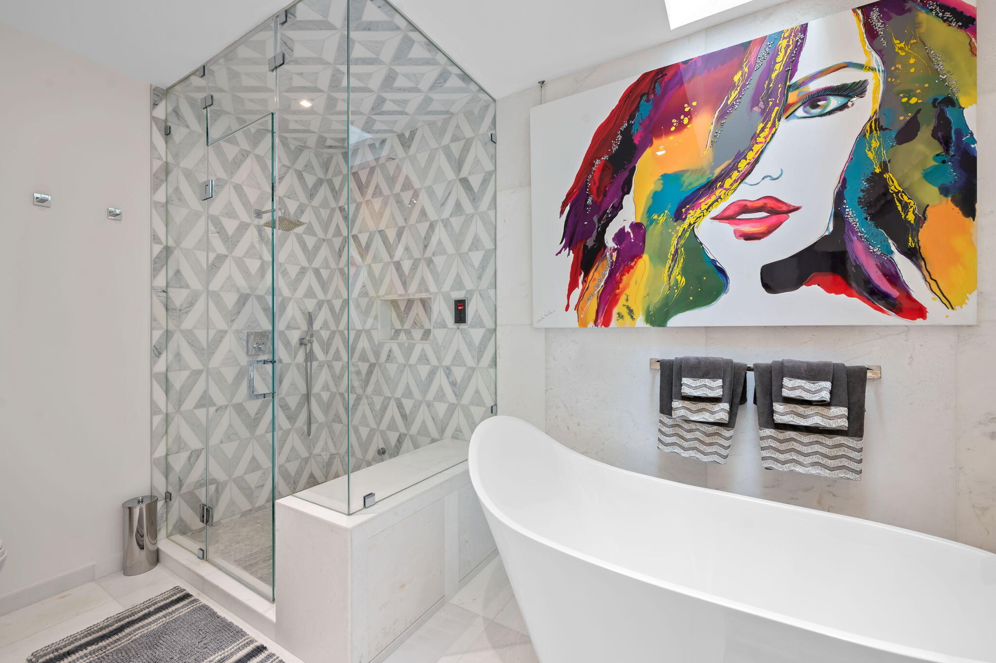 Freestanding soaking tub, glass-enclosed walk-in steam shower with geometric patterned tile, and polished natural stone wall surfacing. Newly remodeled