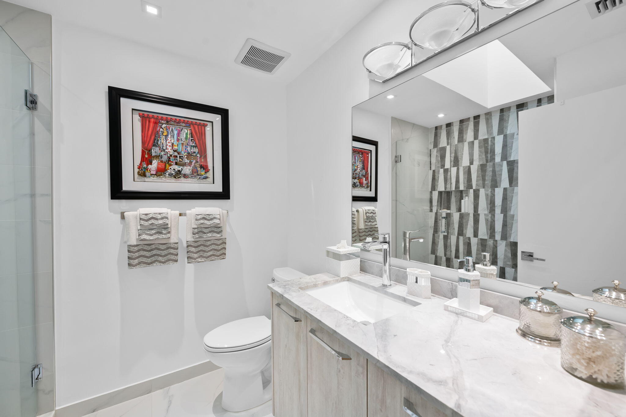 Vanity featuring a light-toned wood-finish cabinet and stone-surface countertop with an integrated basin, large wall-mounted mirror, geometric tile shower surround, and glass-enclosed shower