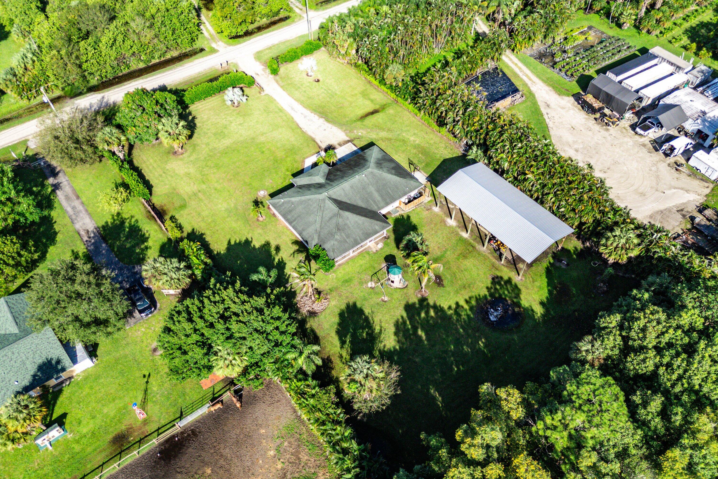 Aerial perspective of a sprawling property featuring a multi-hipped roofline, an expansive lawn, mature palm trees, a detached open-air metal-roof structure, and perimeter fencing