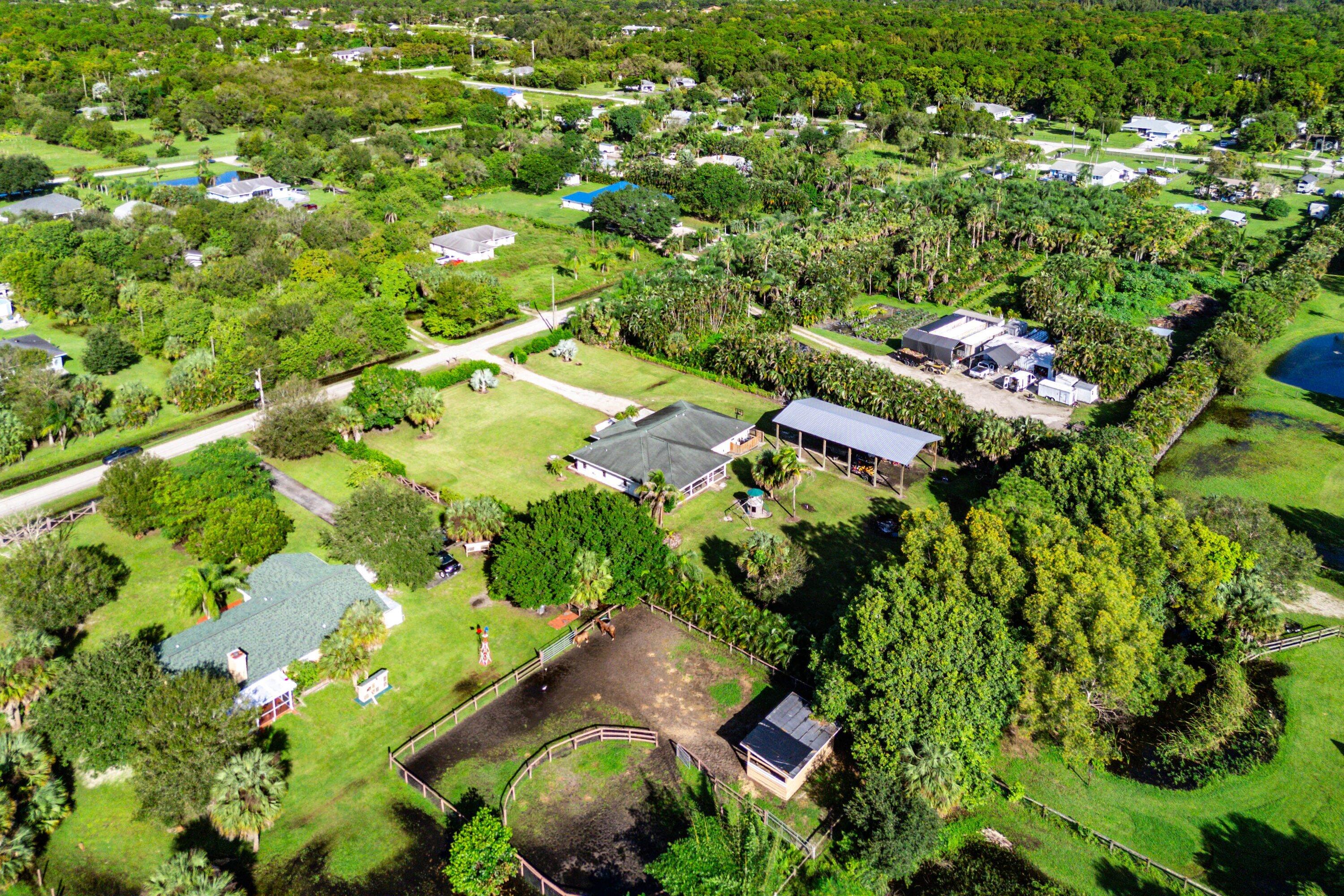 Aerial perspective of a sprawling property featuring a detached metal-roofed structure, fenced corral, mature canopy trees, and an expansive lawn