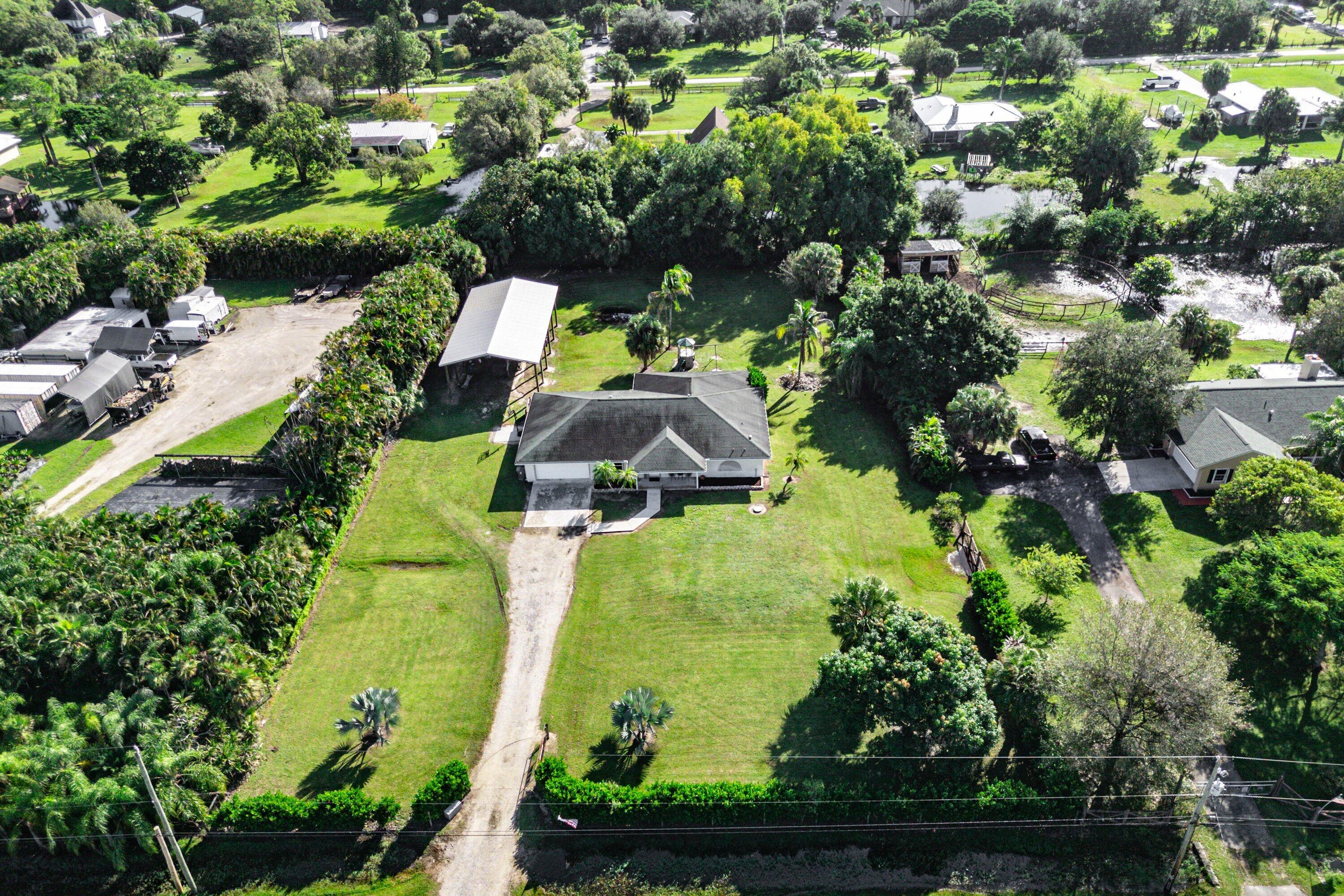 Aerial view of a property featuring a single-story residence, a detached metal-roofed structure, a gravel driveway, and expansive lawn areas with mature trees