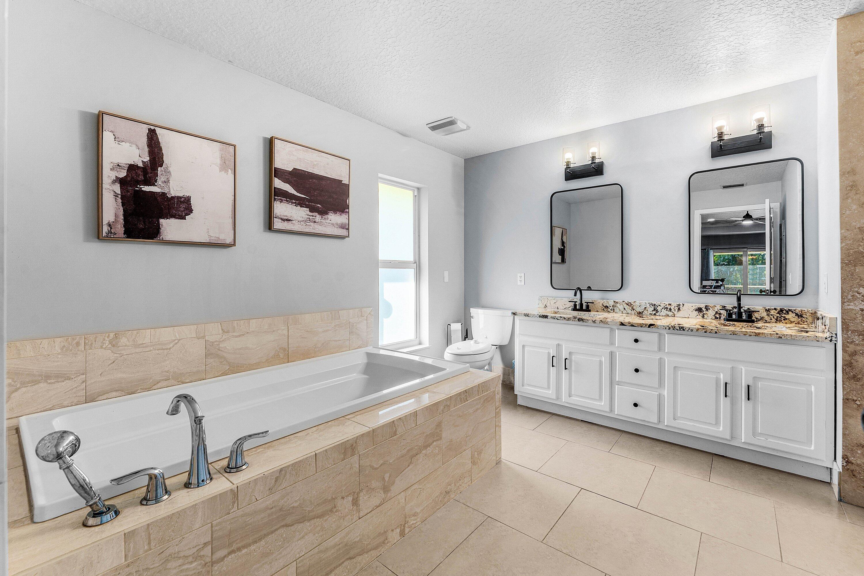 Dual-sink vanity with stone countertops and matte black fixtures, deep soaking tub with tile surround, and large-format tile flooring