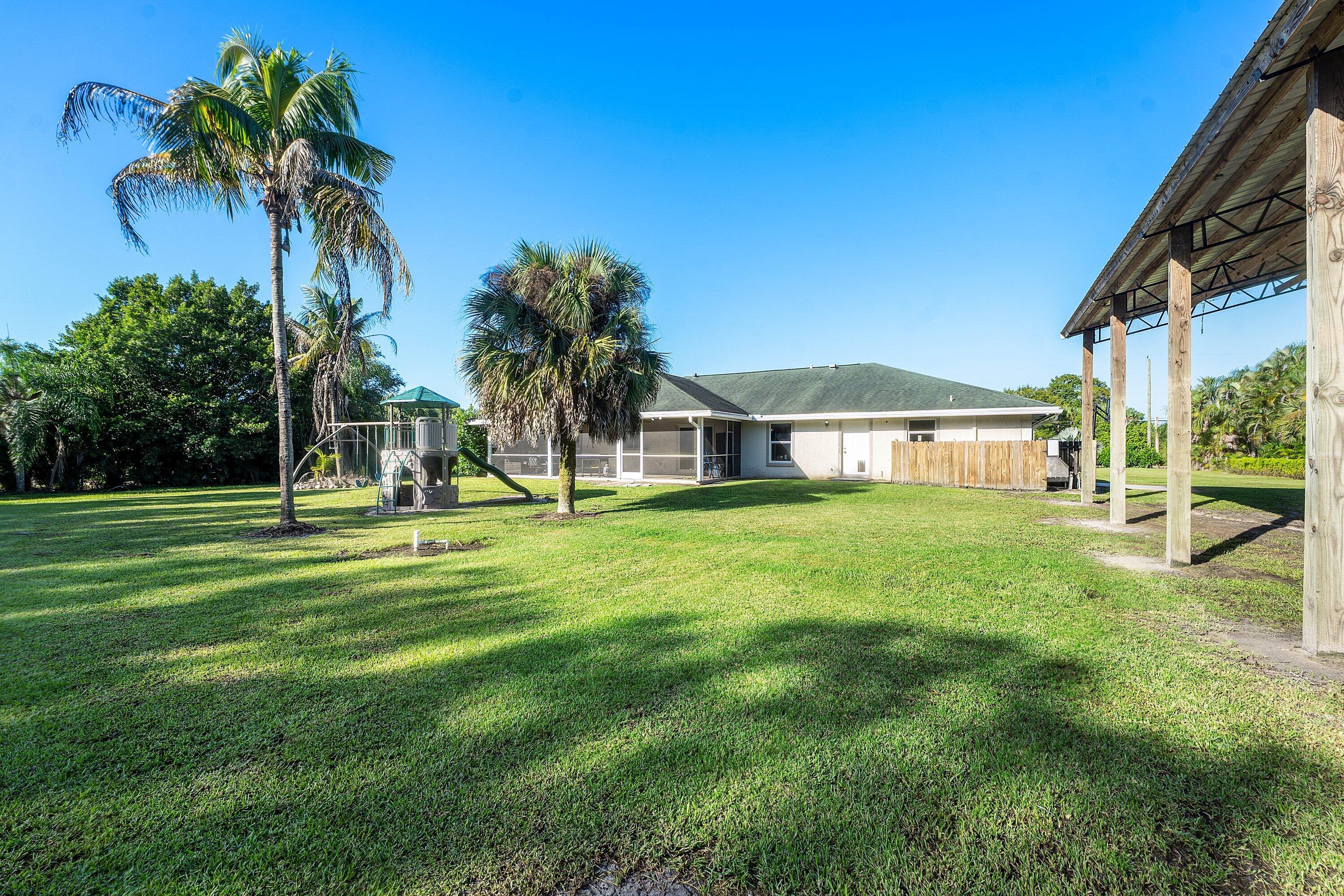 Expansive backyard featuring a sprawling lawn, mature palm trees, a screen-enclosed patio, and a covered timber-frame structure