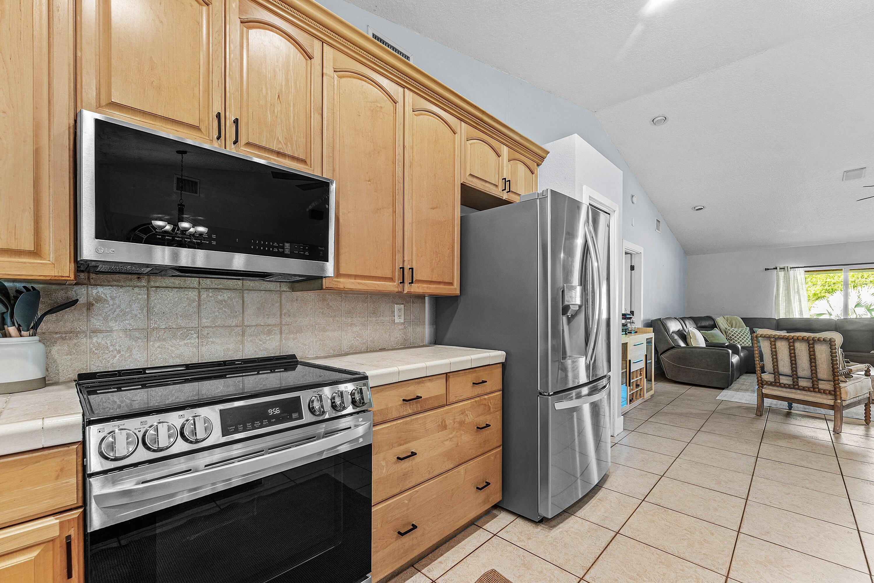 Kitchen featuring natural wood-finish cabinetry, tile countertops, tile backsplash, and stainless-steel appliances including a built-in microwave and refrigerator