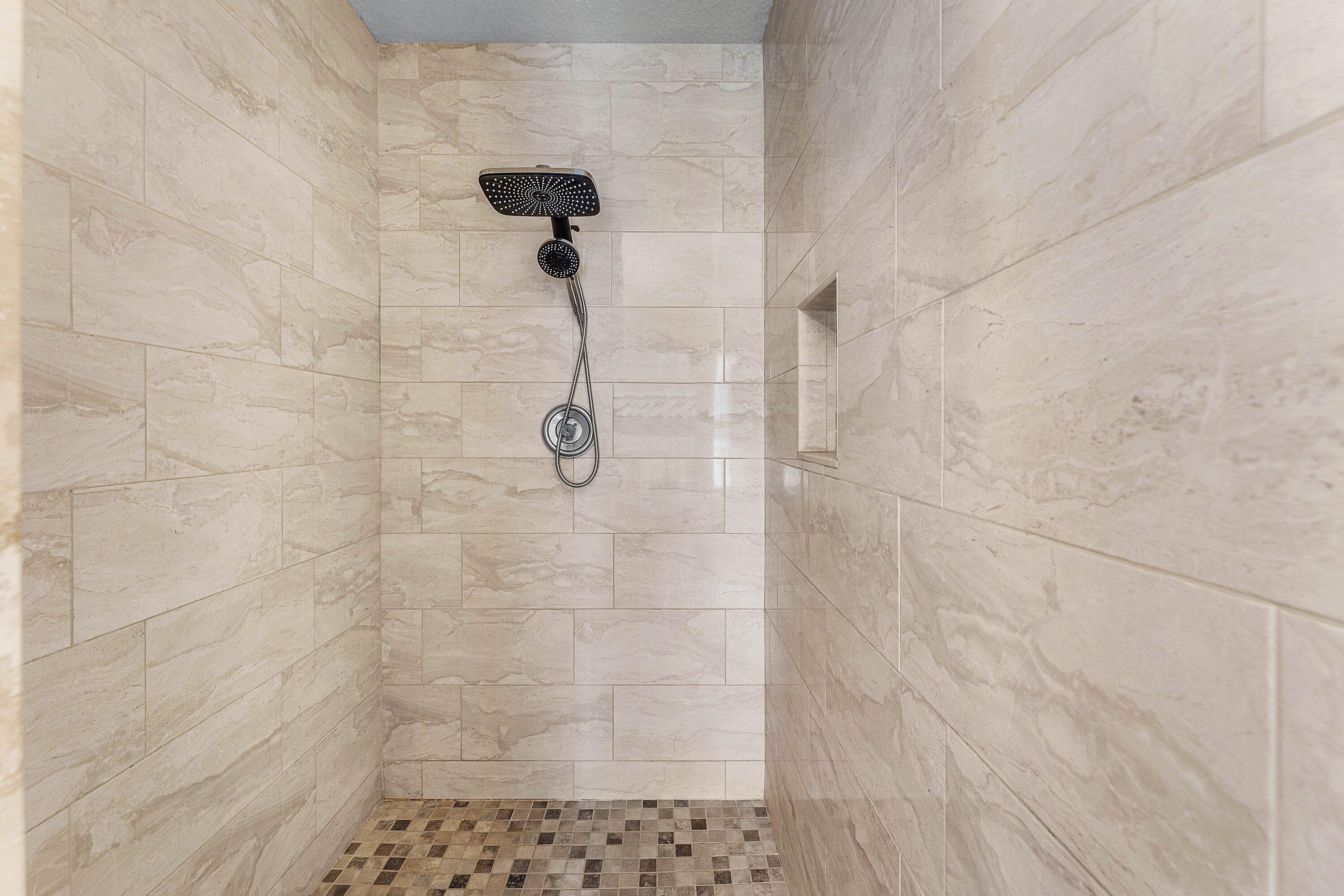 Walk-in shower featuring large-format stone-finish wall tile, a square rainfall showerhead, a hand-held shower sprayer, a built-in recessed wall niche, and mosaic tile flooring