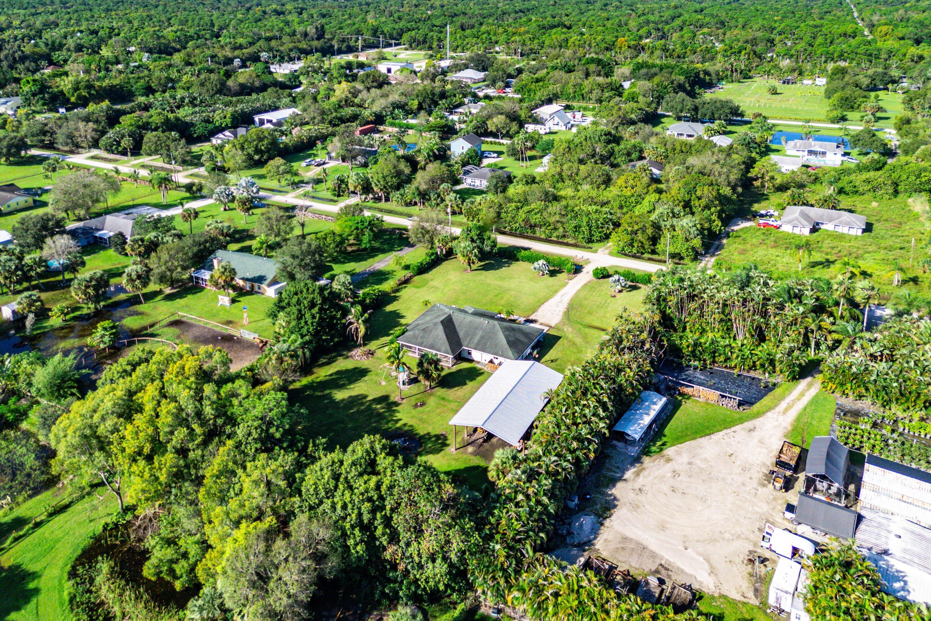 Aerial perspective of a sprawling property featuring a large residence, expansive green space, mature trees, a detached metal-roofed structure, and an unpaved driveway