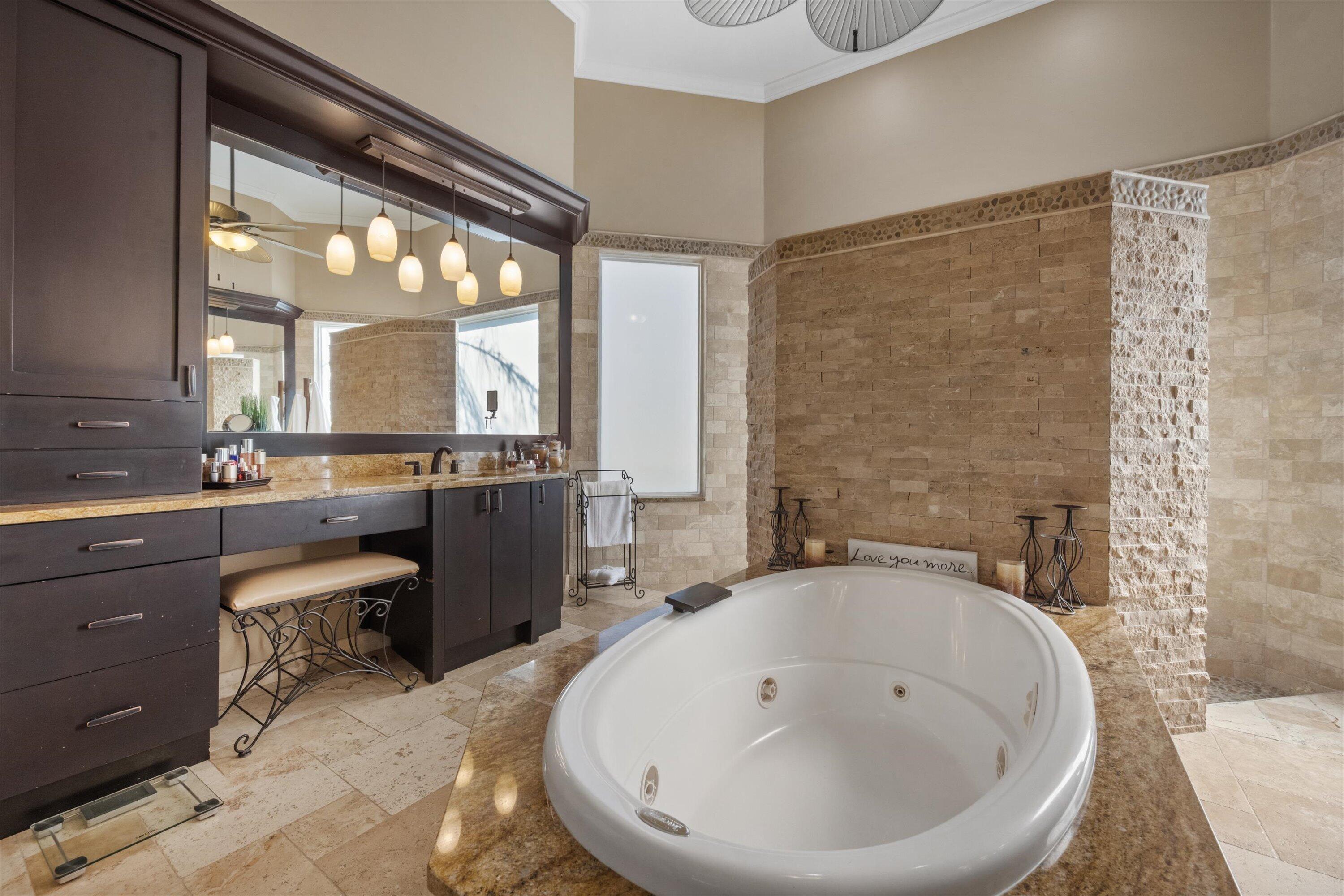 Bathroom featuring a deep soaking tub with a stone-surround deck, stacked stone wall accents, dark wood-finish cabinetry, and a large framed mirror with integrated pendant lighting