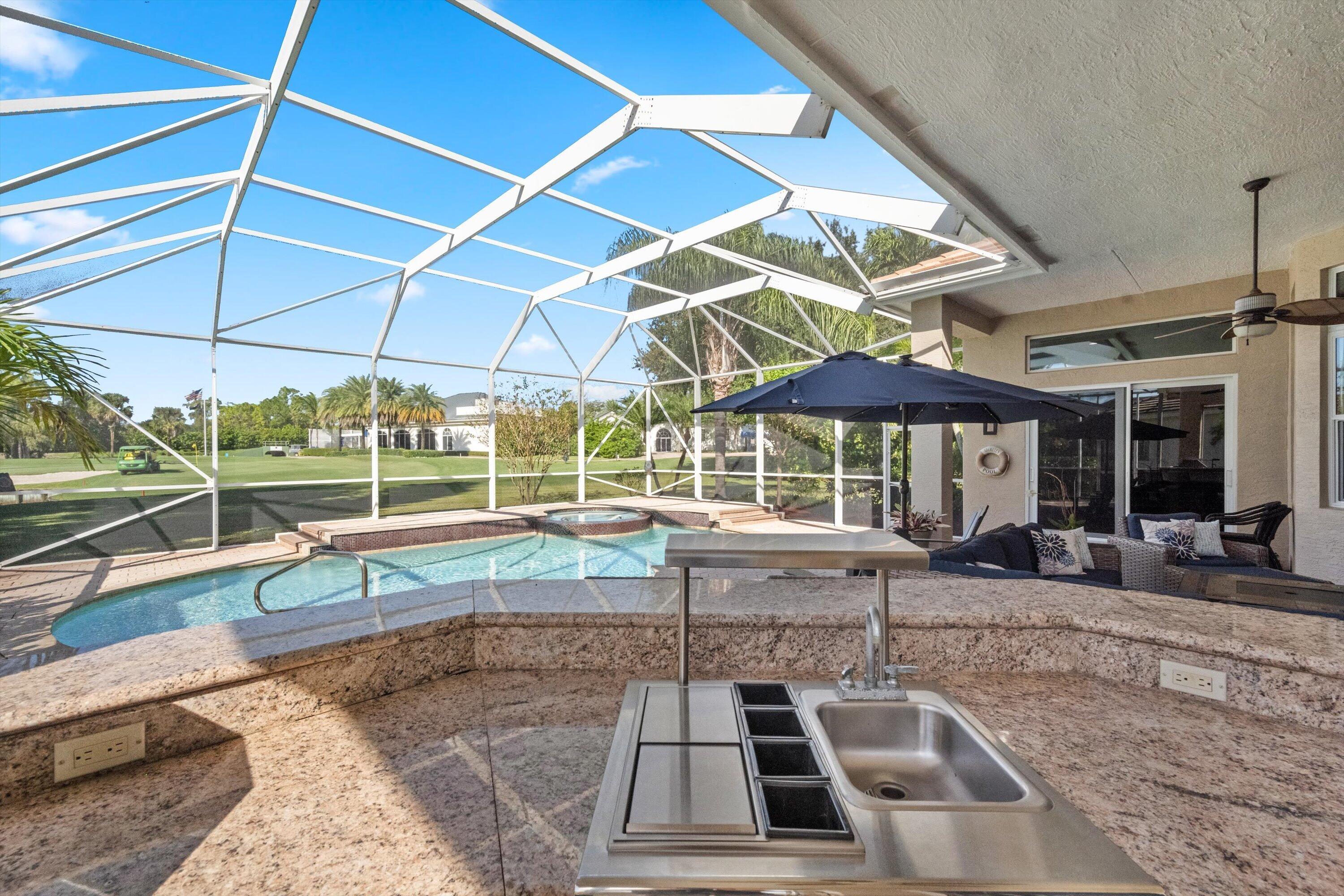 Screened outdoor living area featuring a custom pool, integrated spa, built-in stone-surfaced kitchen island with stainless steel sink, and overhead ceiling fan
