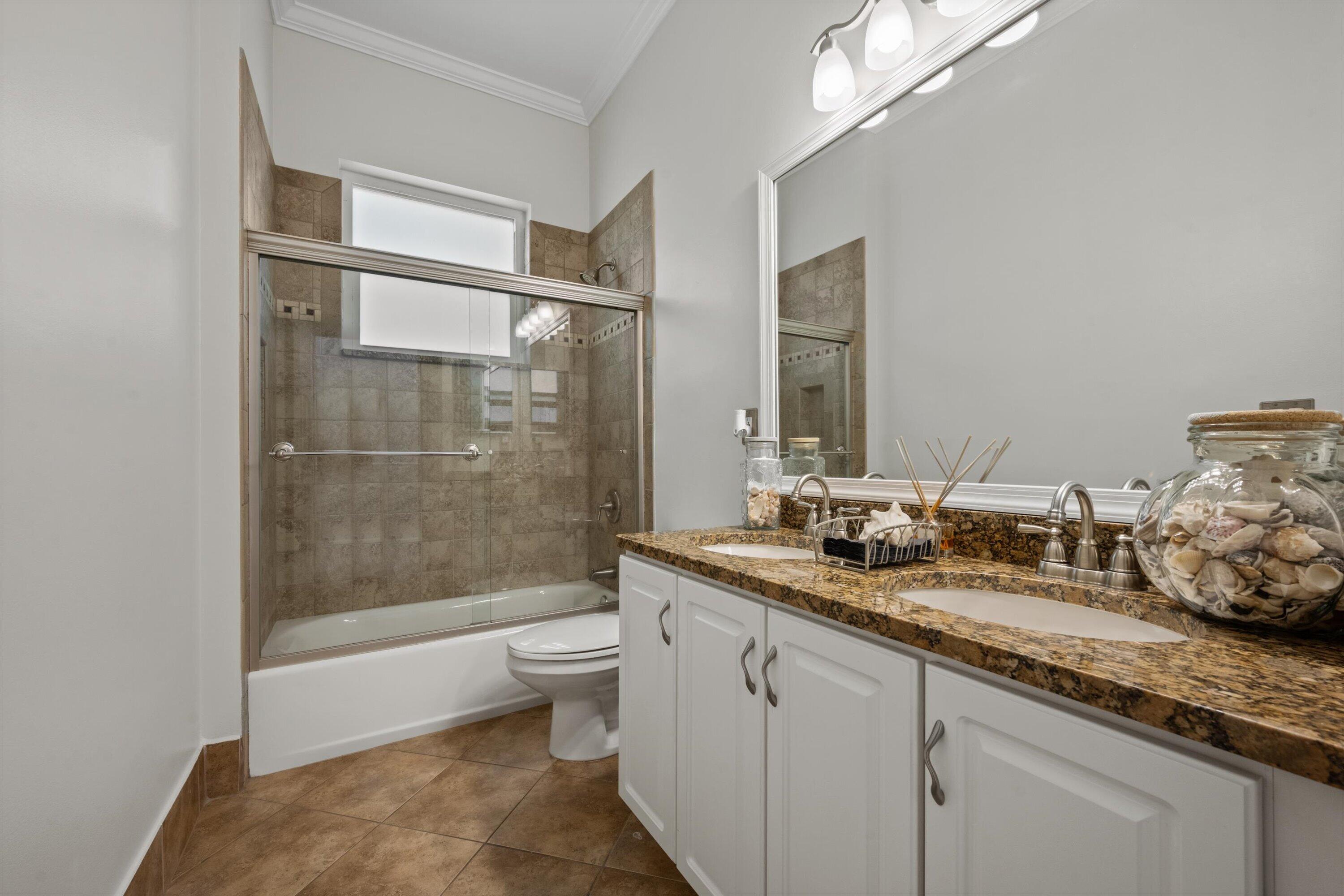 Dual-sink vanity with stone-finish countertop, white shaker-style cabinetry, bathtub with tile surround, glass sliding shower door, and tile flooring