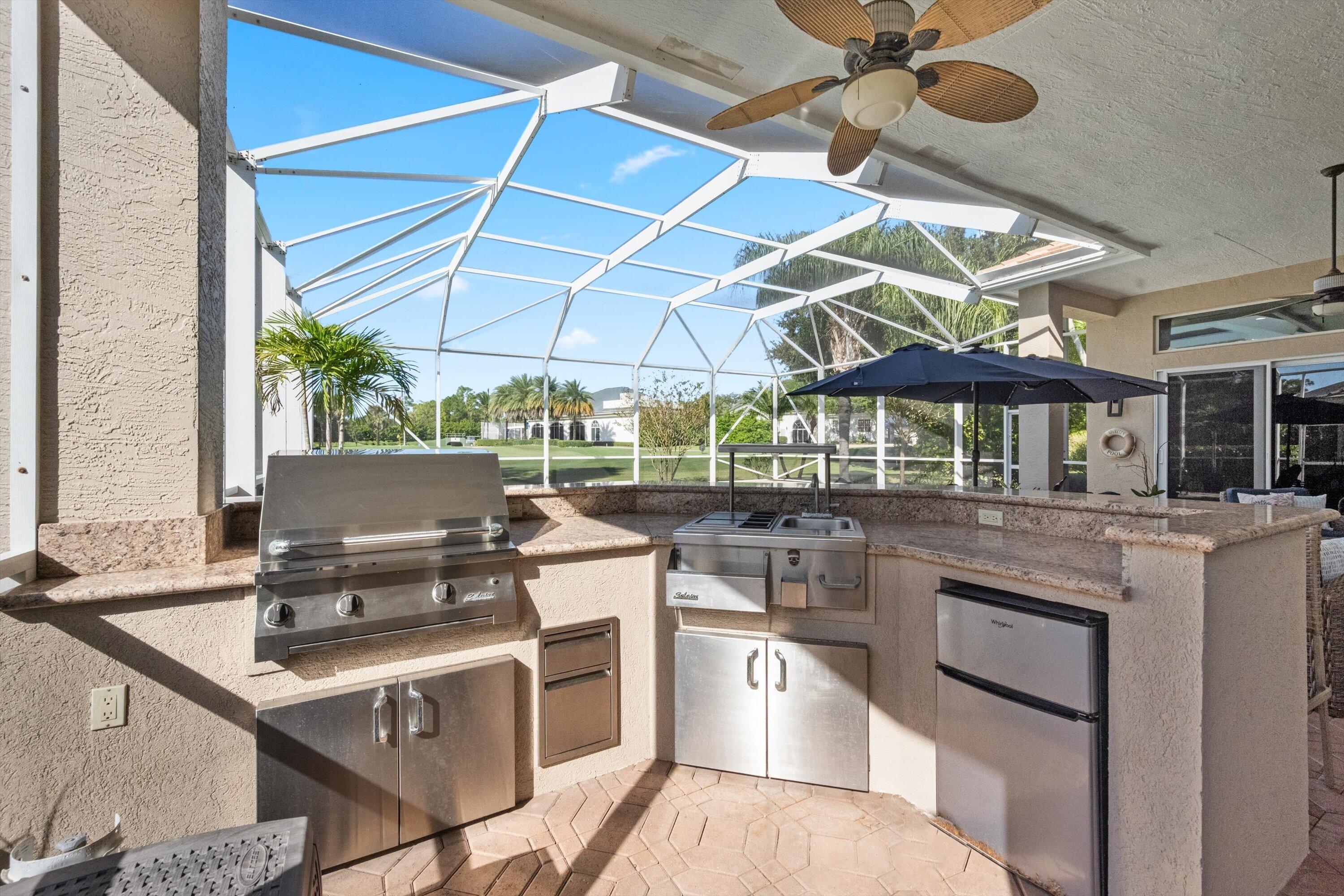 Covered patio featuring an integrated outdoor kitchen with stone countertops, stainless steel grill, stainless steel under-counter refrigerator, and a screened-in enclosure with a ceiling fan