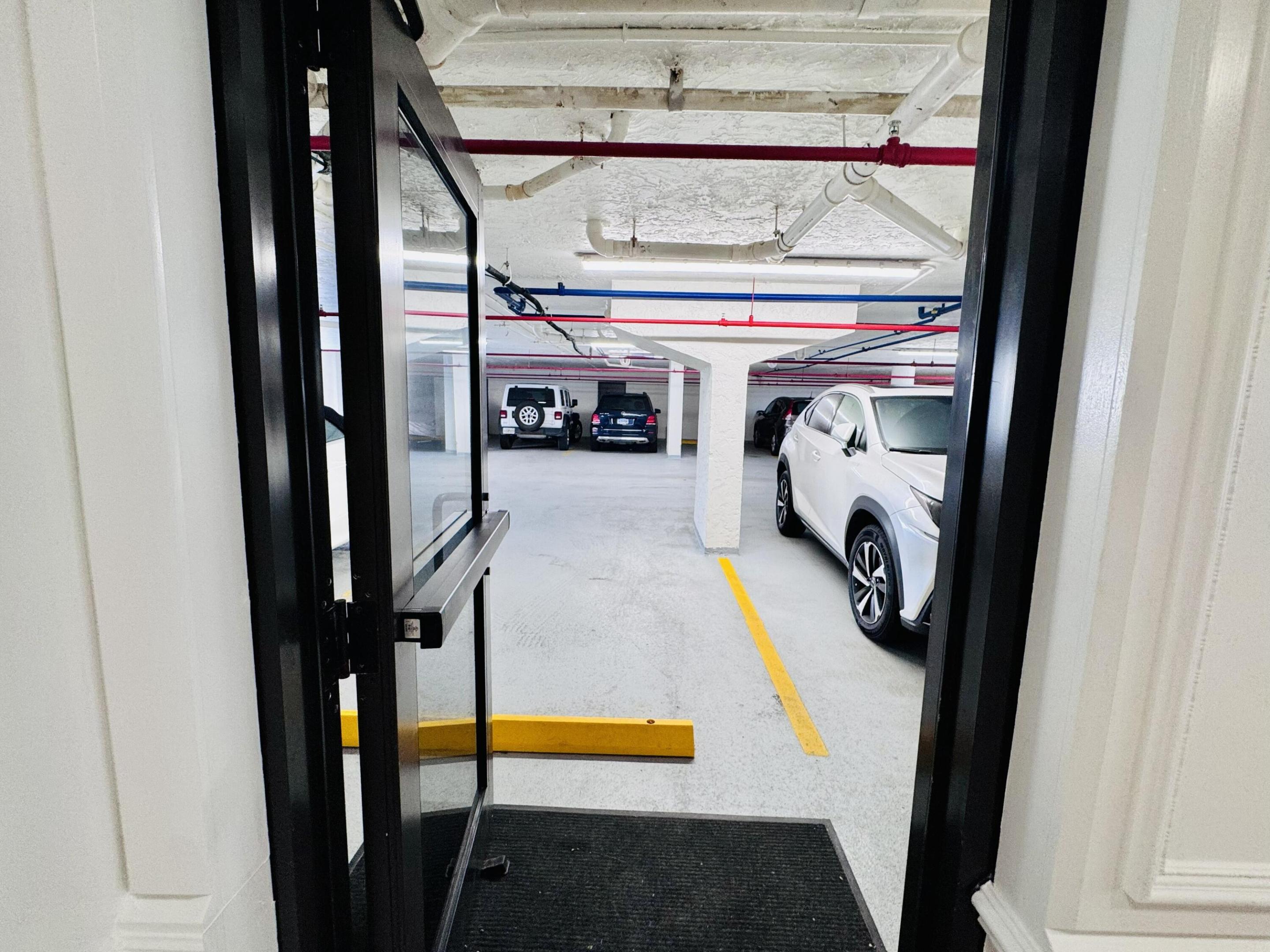 Covered parking area featuring concrete flooring, designated painted parking stalls, structural support columns, and overhead utility piping