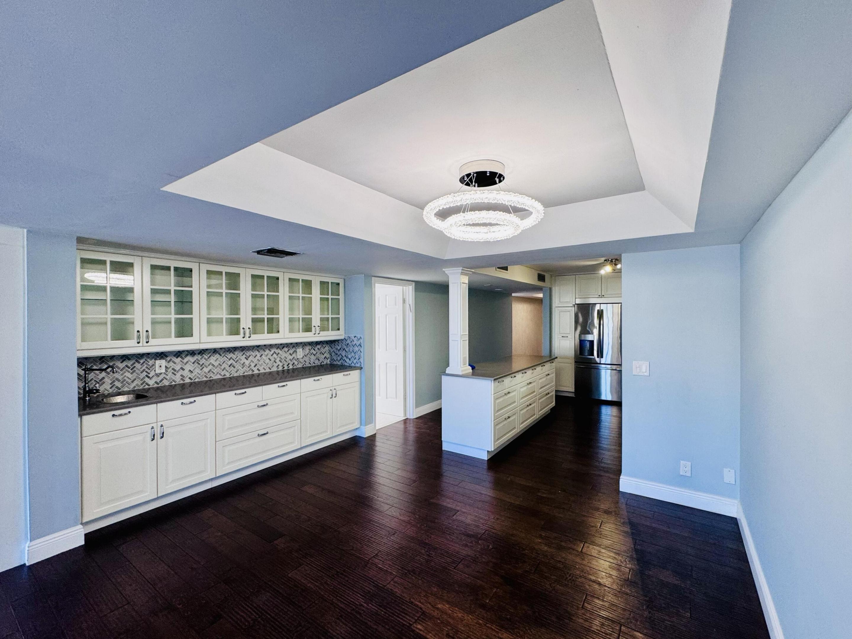 Tray ceiling with integrated circular lighting, wood-finish flooring, custom white cabinetry with glass-front uppers, herringbone-pattern tile backsplash, and dark stone-surface countertops Tray ceiling with integrated circular lighting, wood-finish flooring, custom white cabinetry with glass-front uppers, herringbone-pattern tile backsplash, and dark stone-surface countertops