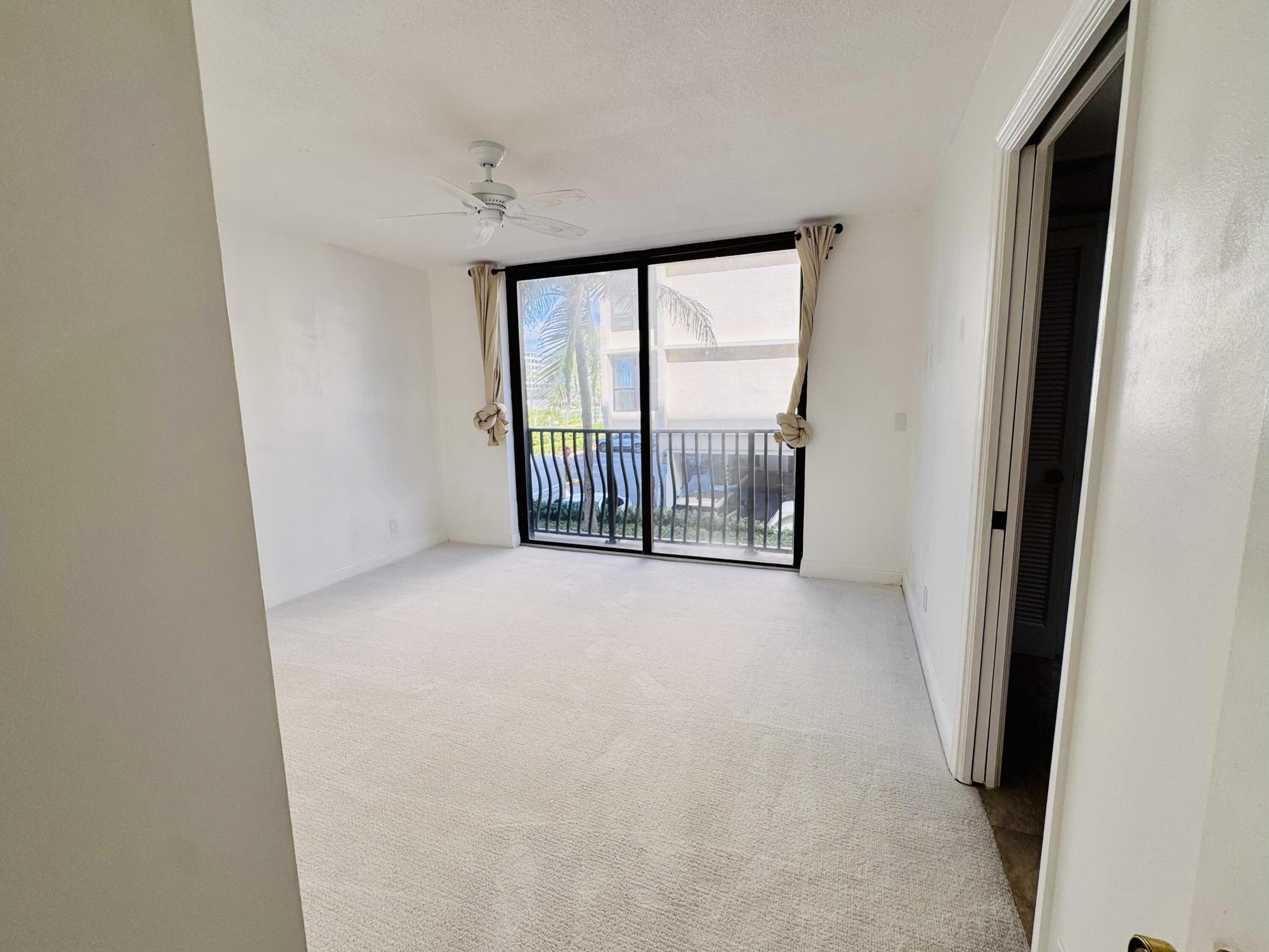 Neutral-toned wall-to-wall carpeting, ceiling-mounted paddle fan, and large floor-to-ceiling glass sliding doors opening to a private balcony with metal railing