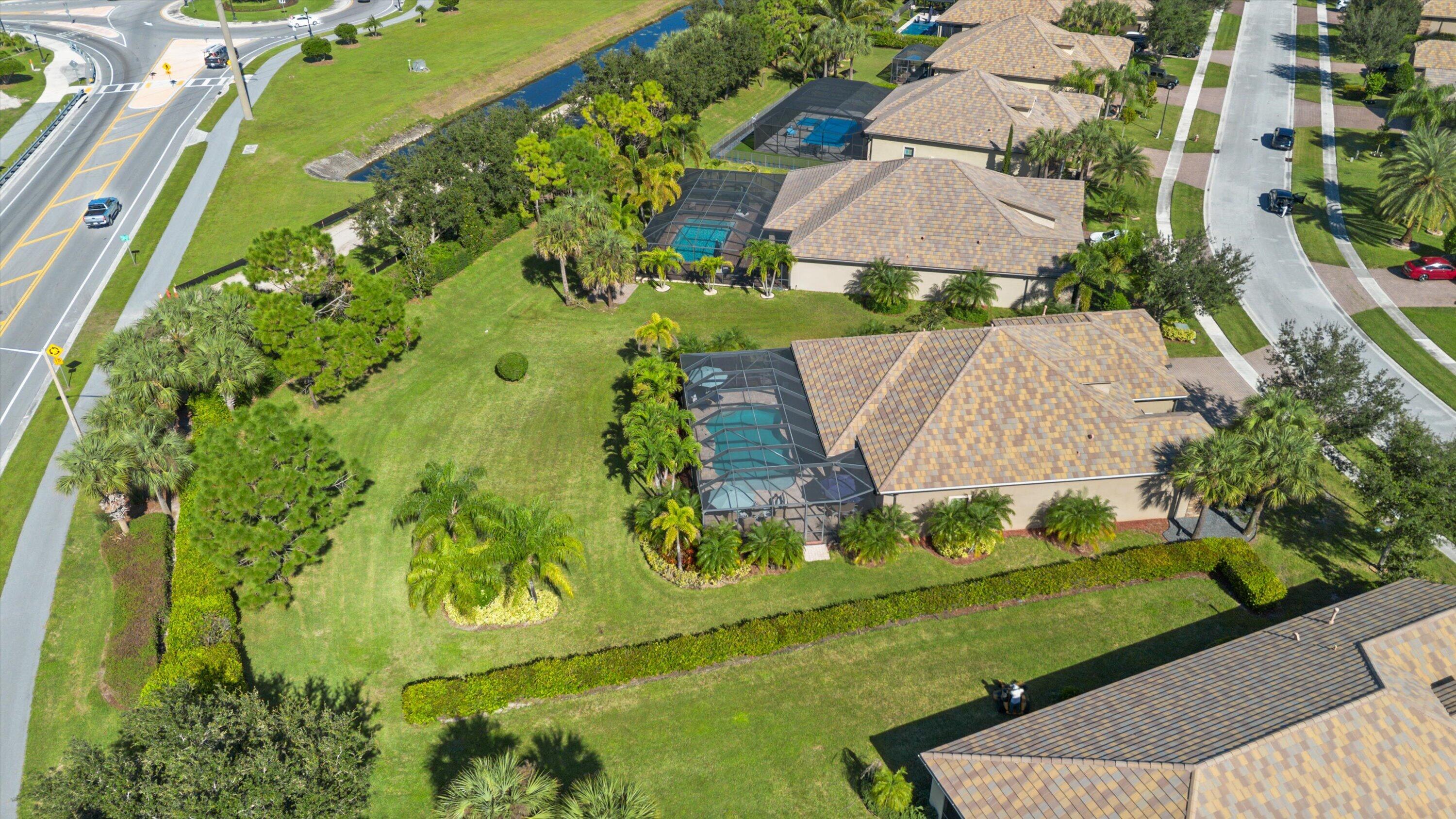 Aerial view of property featuring a screened-in swimming pool, shingle roof, expansive lawn, mature palm trees, and perimeter landscaping
