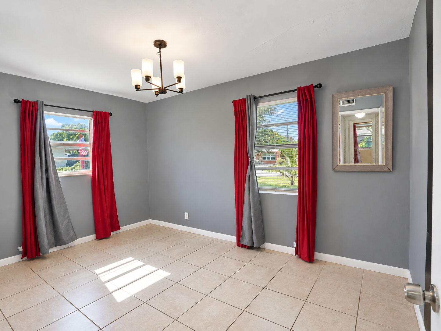 Neutral-toned interior space featuring light-colored ceramic tile flooring, dual windows with black curtain rods, and a multi-arm modern chandelier