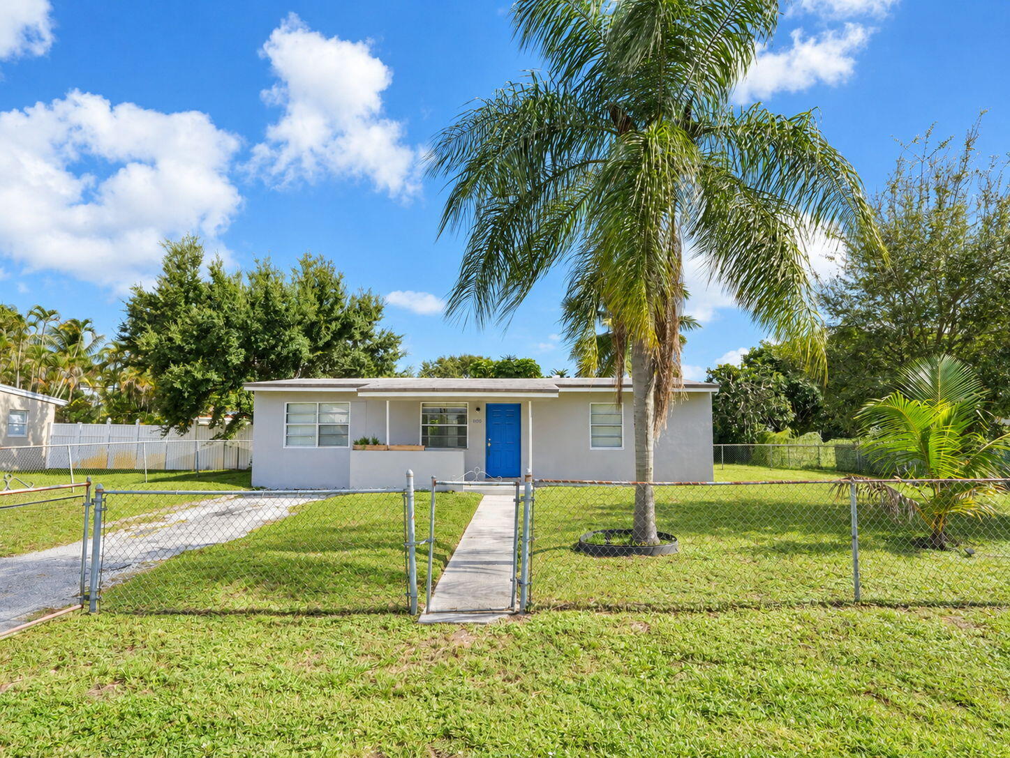 Single-story exterior featuring a concrete walkway, chain-link perimeter fencing, and mature palm landscaping Single-story exterior featuring a concrete walkway, chain-link perimeter fencing, and mature palm landscaping