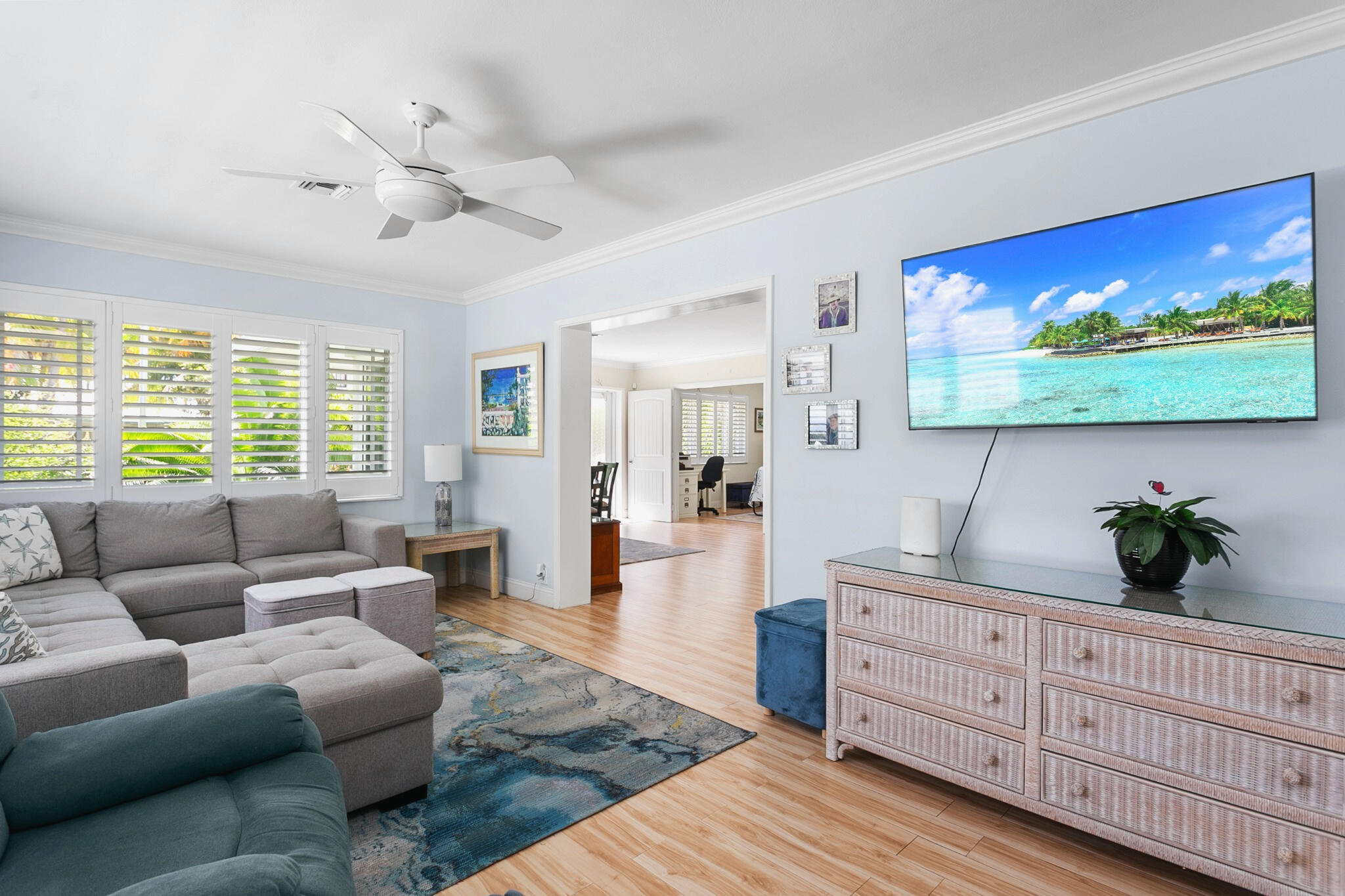 Living area featuring light-toned wood-finish flooring, white plantation shutters, crown molding, and big picture windows allowing lots of natural light