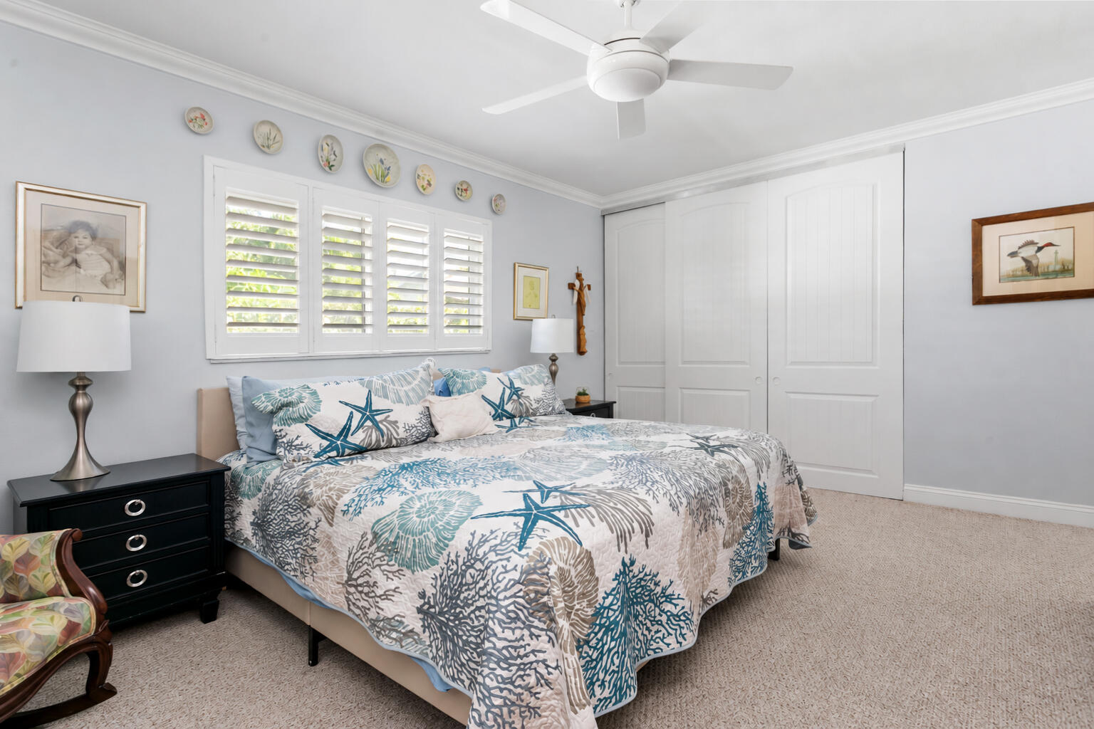 Bedroom featuring white plantation shutters, neutral carpeting, ceiling fan, and floor-to-ceiling sliding closet doors with paneled detailing