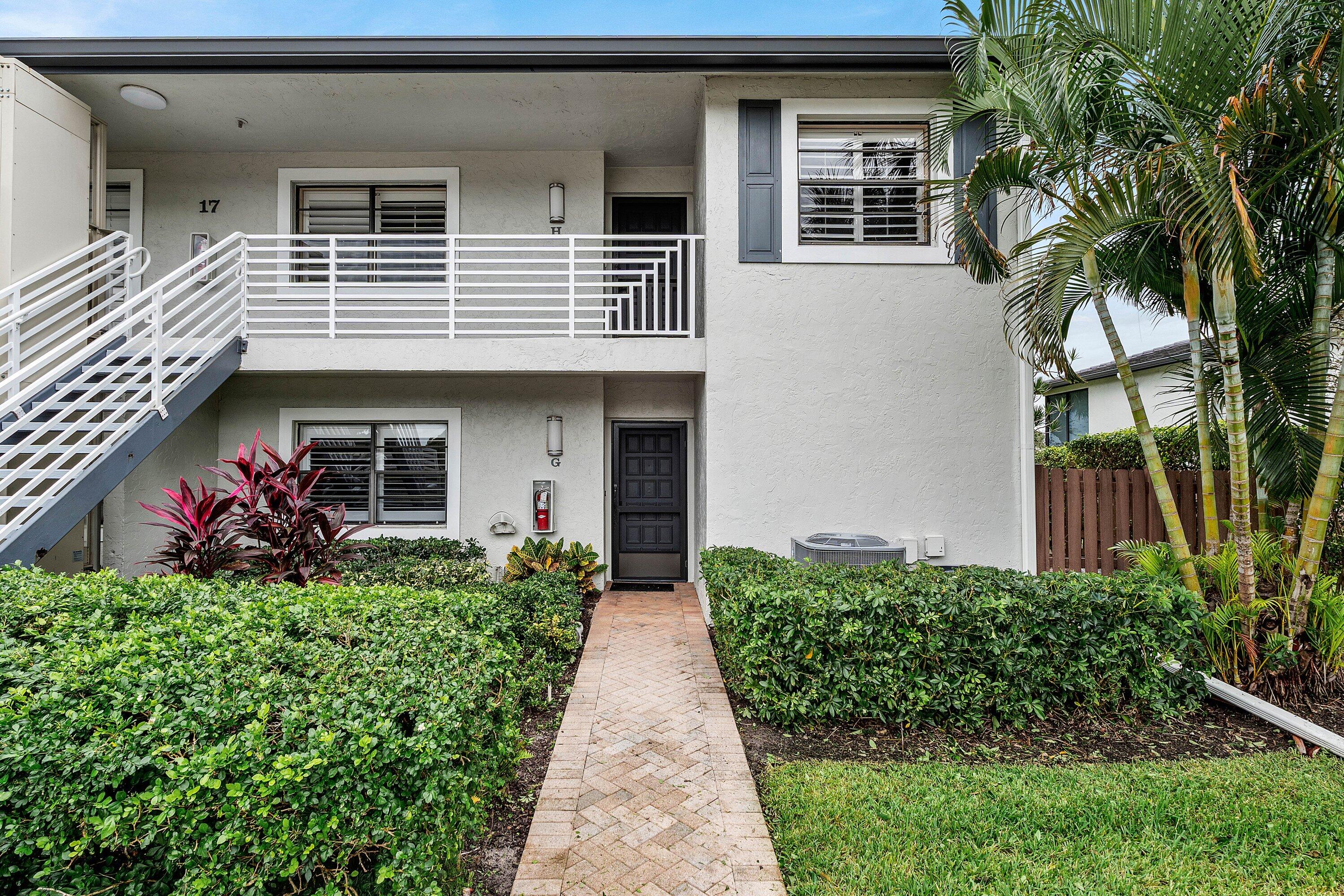 Two-story building exterior featuring a textured stucco facade, a brick-paved walkway, a second-level balcony with white horizontal railings, and mature landscaping