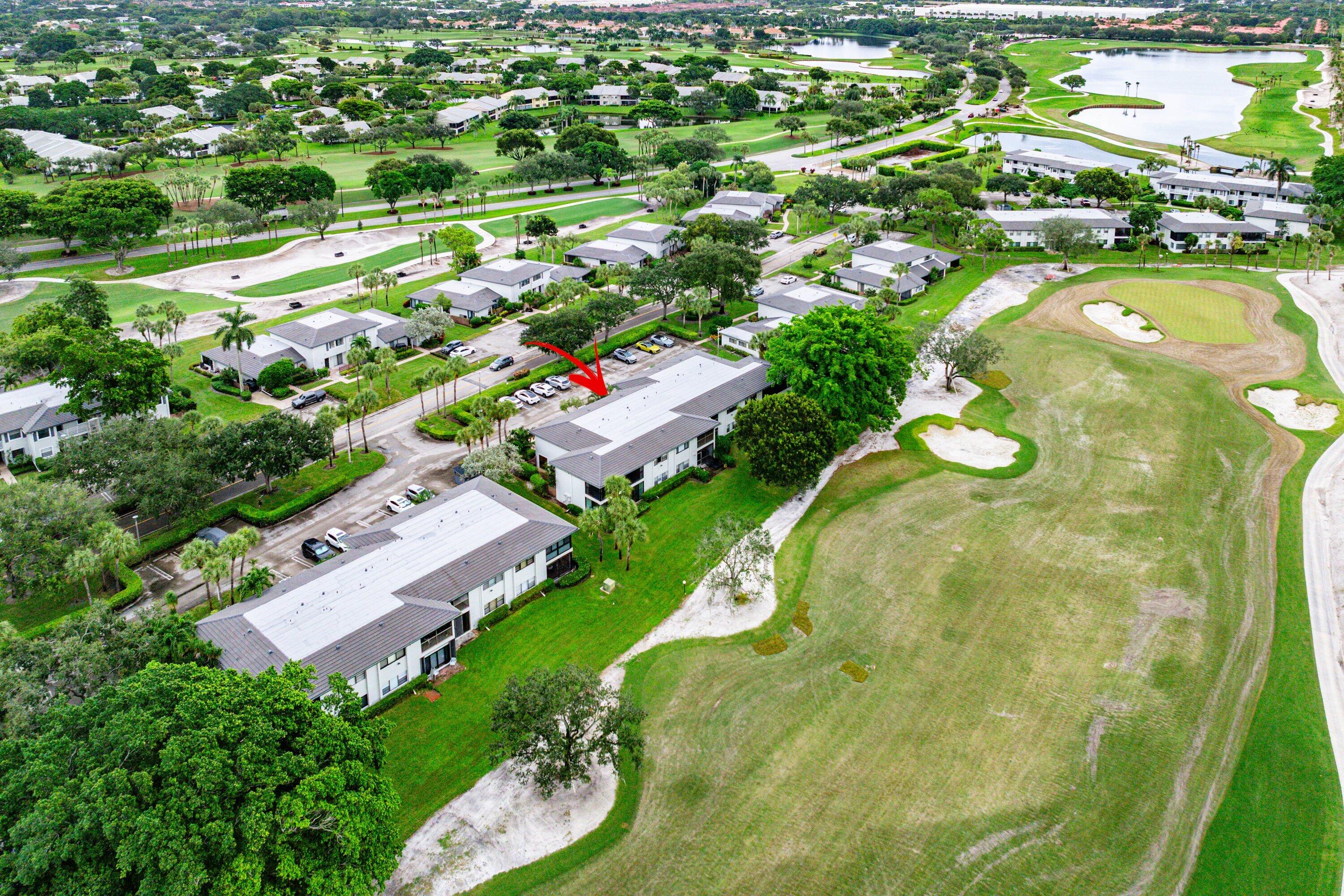 Aerial perspective of a multi-unit residential building featuring a light-toned roof, surface parking area, mature landscape vegetation, and proximity to an expansive golf course with water features